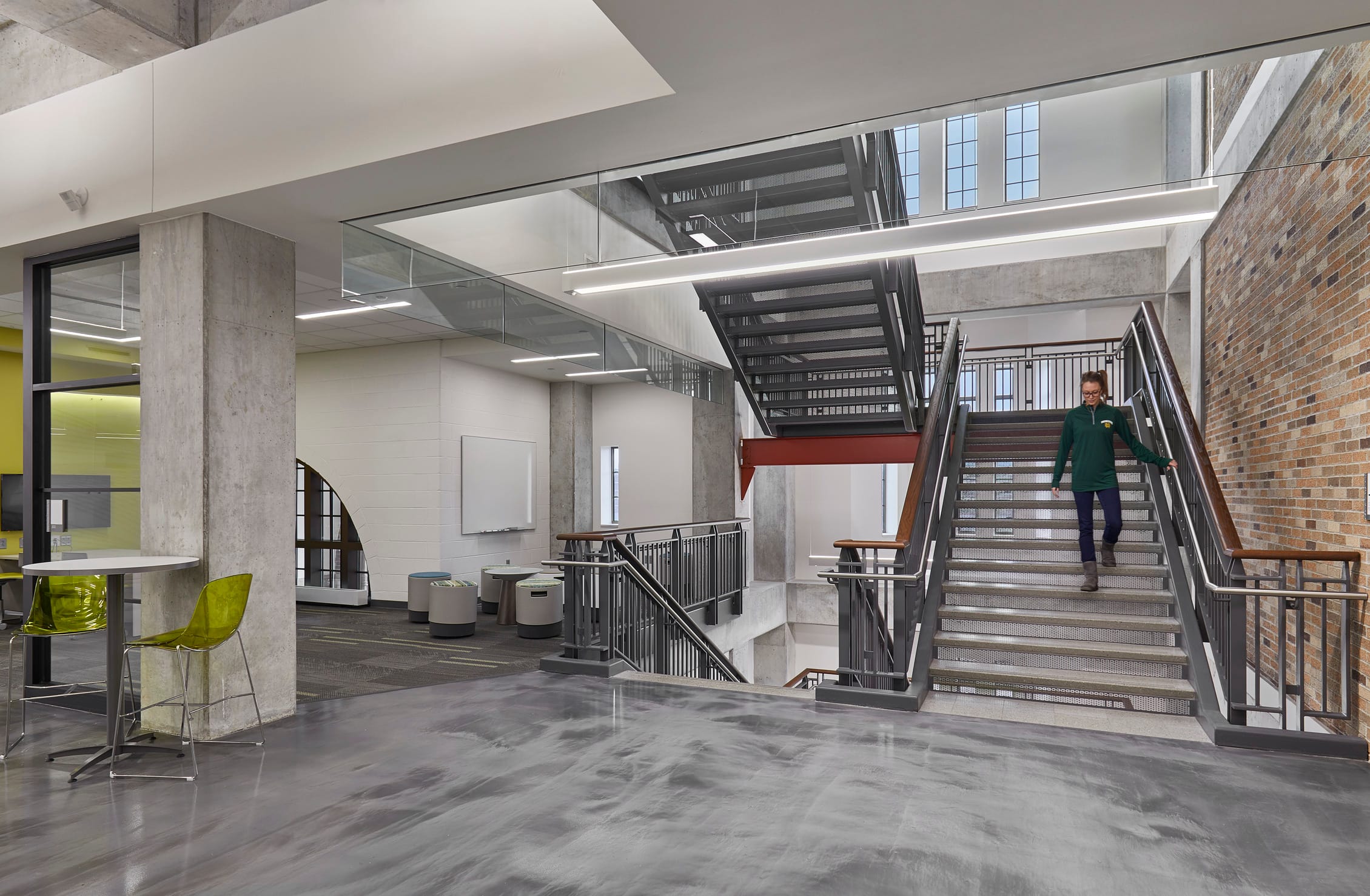 This image shows a modern, open-concept academic space featuring contemporary design elements. This image specifically is focusing on the architectural elements of a metal staircase with red accents against a red brick wall. The space is a multi-story atrium with exposed concrete structural elements like exposed concrete pillars, white brick walls, and exposed brick. The overall design combines industrial elements with bright pops of color in the furniture (like a set of lime green chairs in the foreground) to create an inviting, collaborative environment typical of modern university buildings or innovation centers.