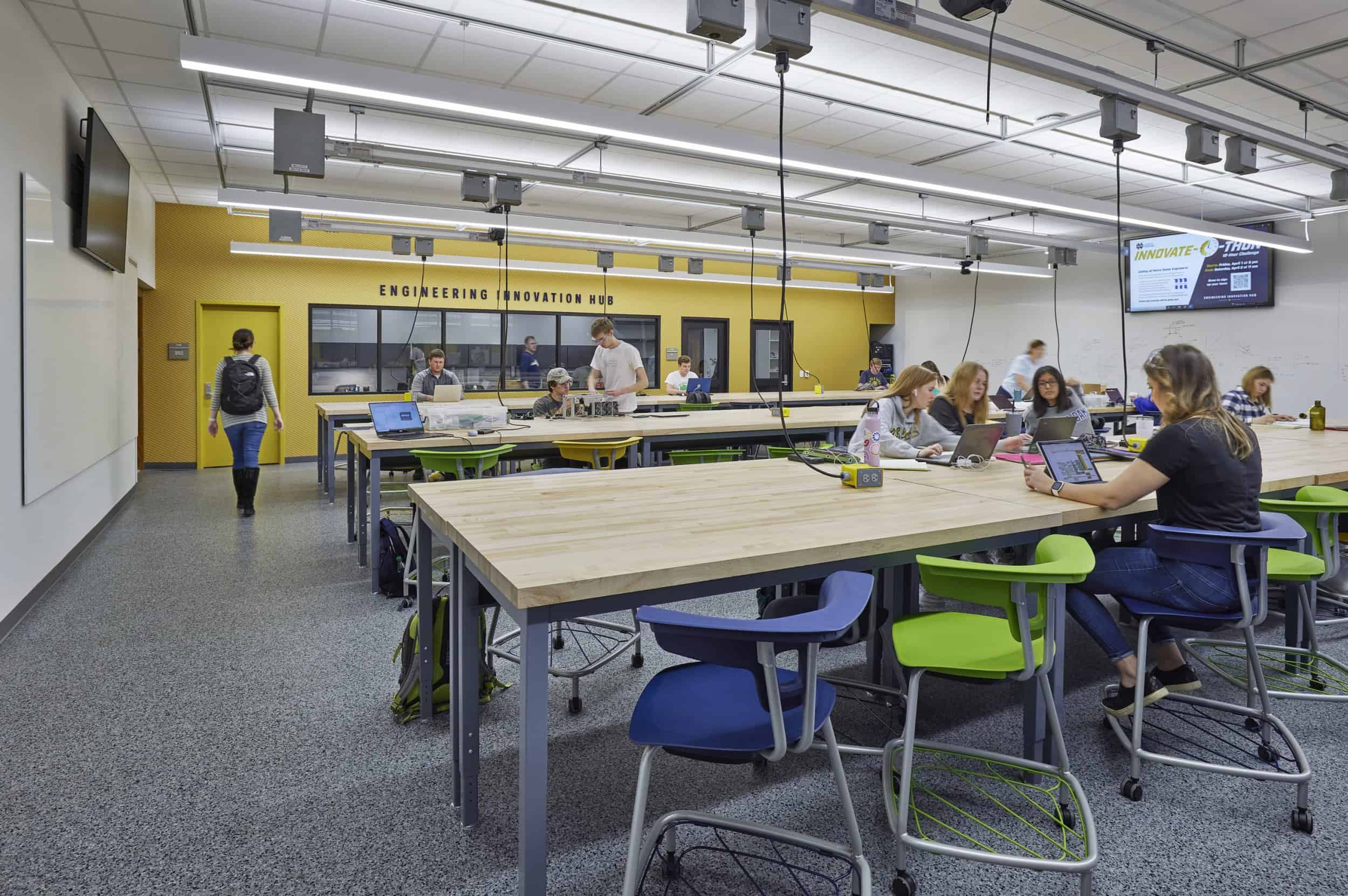 This image shows a modern collaborative workspace or educational makerspace with large wooden worktables and bright yellow chairs. Several people are seated at the tables working on laptops or projects, while a few individuals are walking through the space. The room features blue walls, a white drop ceiling with hanging electrical outlets, and display screens mounted on the walls. The bright, open environment appears designed for group work, with power cords hanging from ceiling tracks to provide electricity to the work areas. The space combines functionality with vibrant colors, creating an energetic atmosphere for creativity and collaboration. In the background 