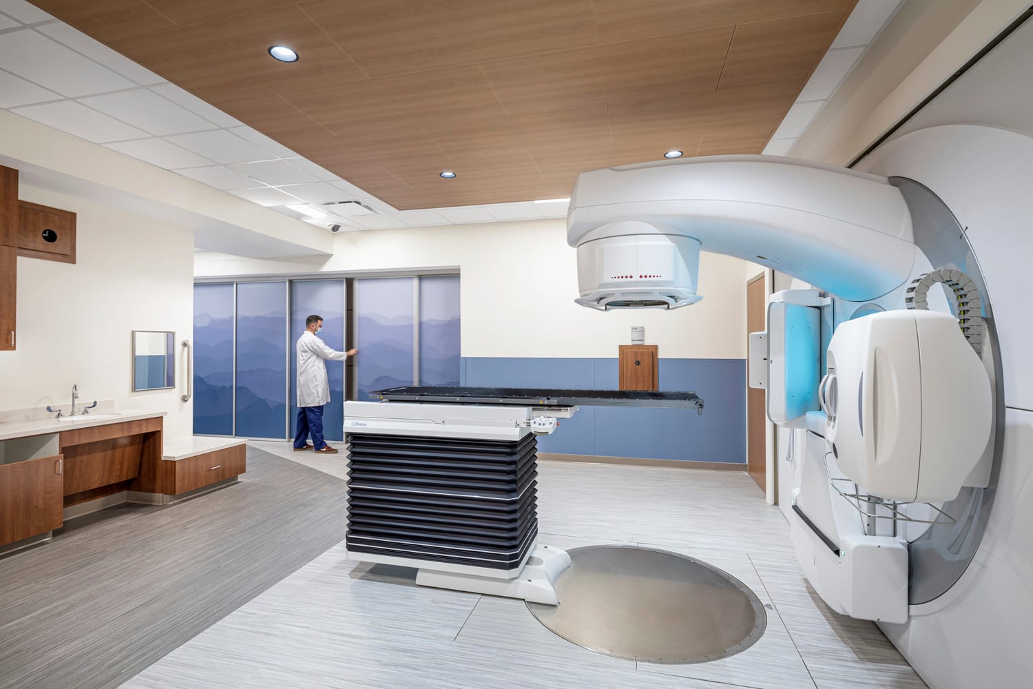 Medical radiation therapy room with advanced linear accelerator equipment. A healthcare professional in a white lab coat stands near sliding glass doors with a mountain landscape view. The room features a large white radiation therapy machine on the right, a patient positioning table in the center, and wood-toned cabinetry and counters on the left. The space has a clean, modern design with white and blue color tones, recessed lighting, and a wood-paneled ceiling.