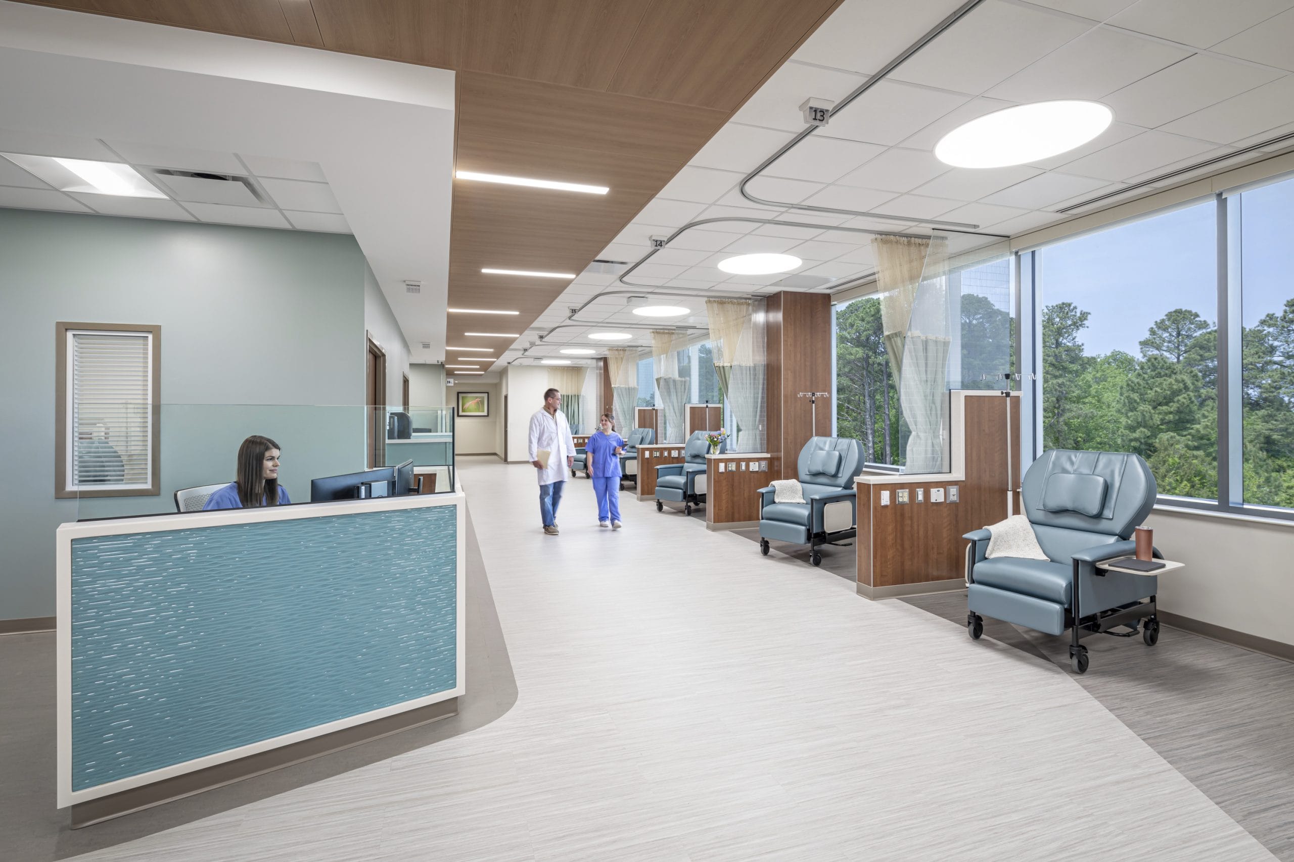 Infusion center or treatment room in the UNC Rex Cancer Center with multiple stations along a floor-to-ceiling windowed wall with a view of lush greenery. Opposite to the infusion chairs is a nurse's station with textured blue siding that looks like ocean waves. Each station has aqua blue reclining chairs is separated by wood partitions.