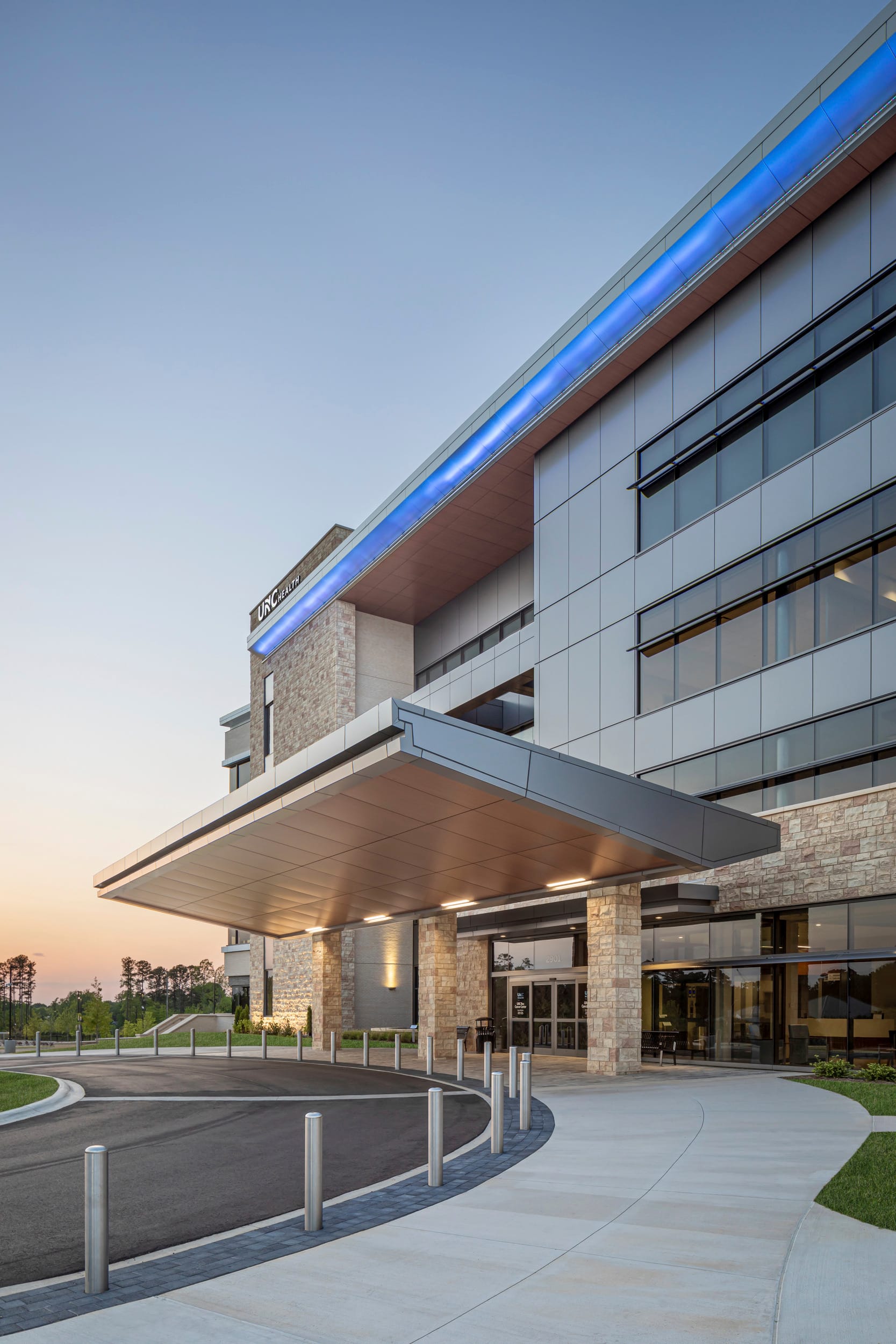 The image shows a modern healthcare facility, specifically a UNC Health Cancer Care center, photographed at dusk or early evening. The multi-story building features a contemporary architectural design with a combination of glass, stone, and metal elements. The front entrance is covered by an extended overhang that looks like metal. The top of the building has a large LED blue light on the roofline, giving the building a distinct blue glow.