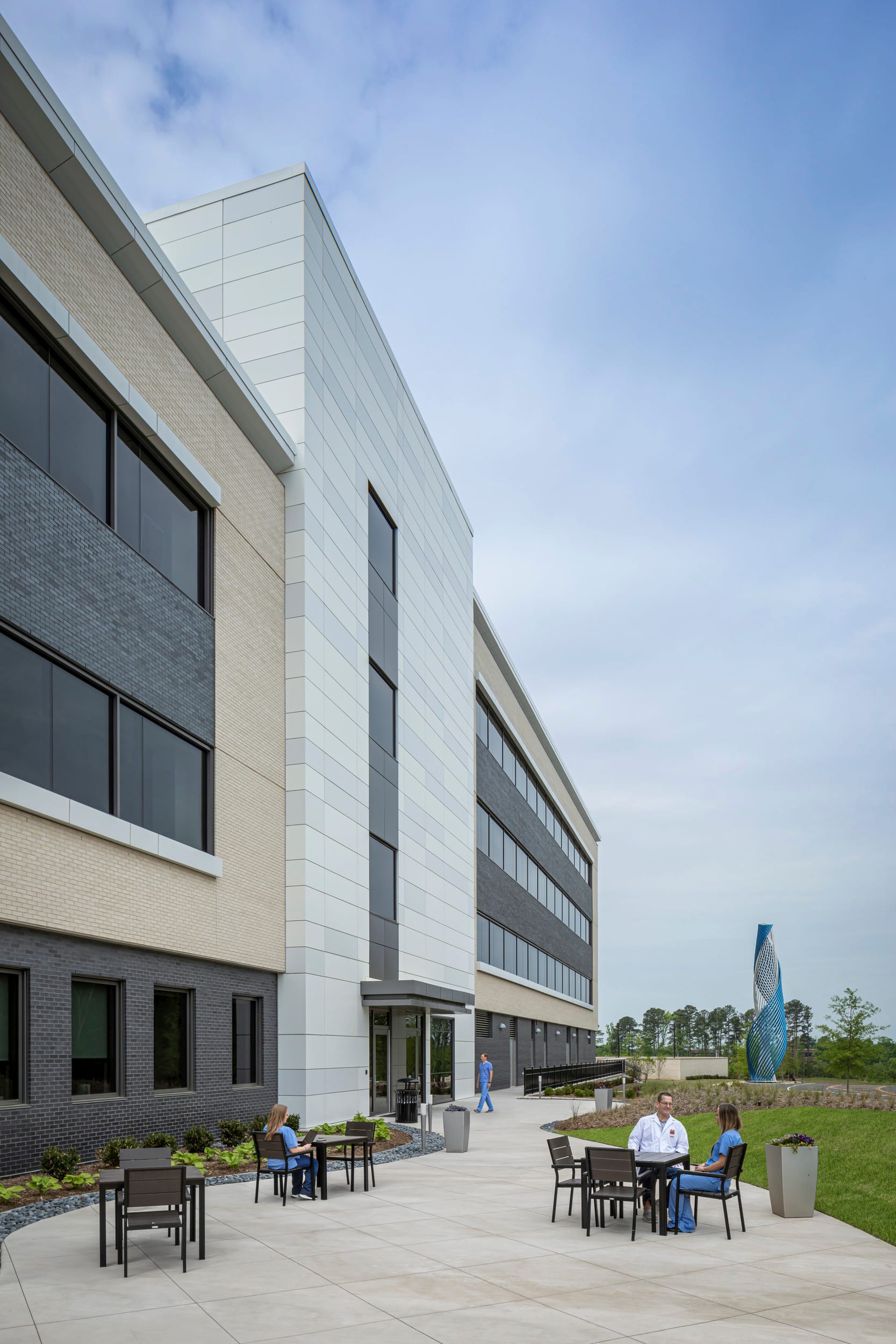 The image shows a close up of a modern healthcare facility, specifically a UNC Health Cancer Care center. The multi-story building features a contemporary architectural design with a combination of glass, stone (specifically, beige and black brick), and metal elements. The landscaping curved walkways leading to the building. There are outdoor tables with chairs where hospital staff are enjoying lunch. The overall design blends functionality with a welcoming aesthetic appropriate for a medical facility.