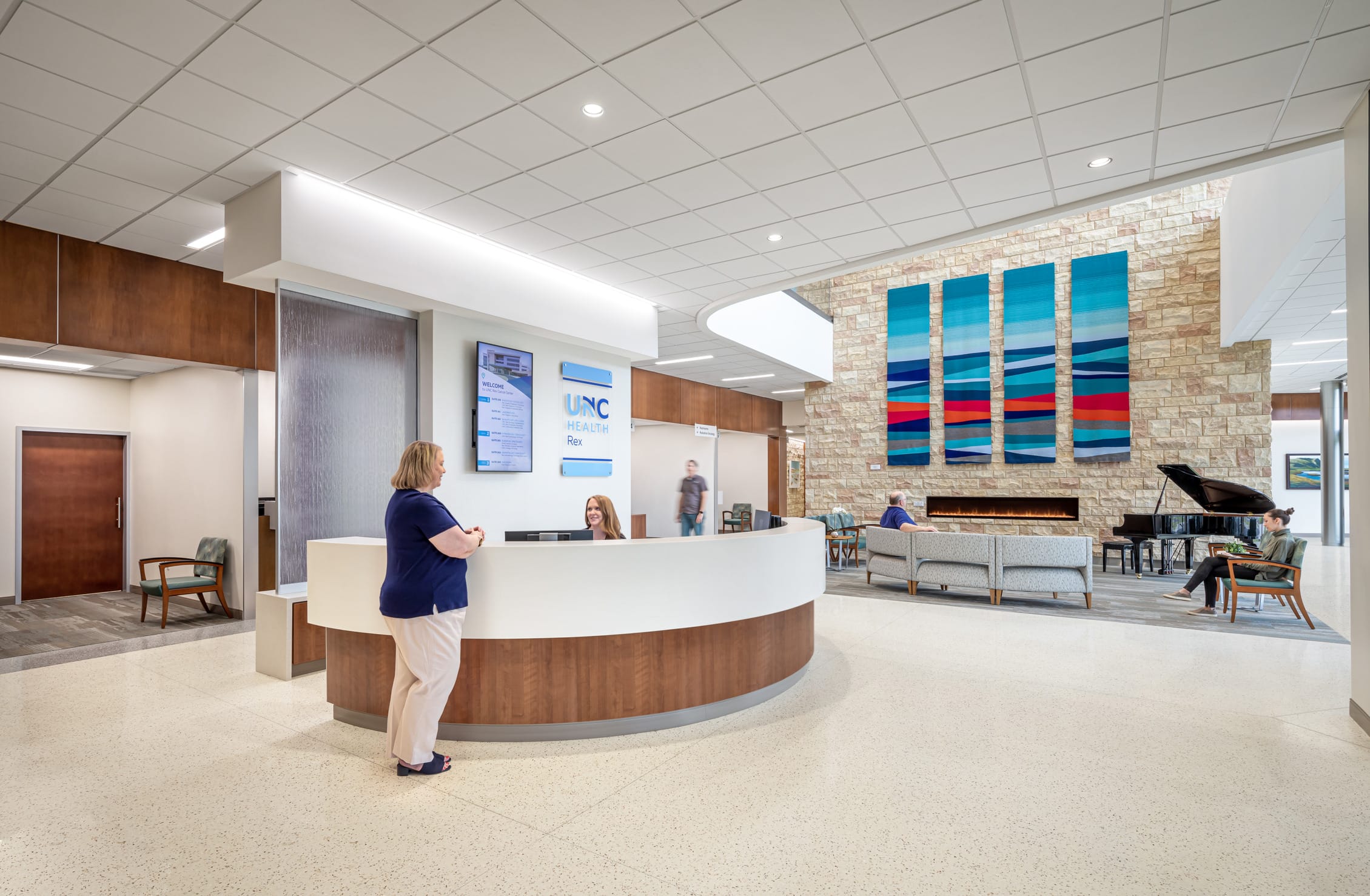 Modern healthcare reception area with a curved white and wood-toned welcome desk. A woman in a navy blue top stands near the desk, with a receptionist seated behind. The space features a stone accent wall with colorful abstract artwork and a linear fireplace. Digital signage displays 'UNC' branding. A grand piano is visible in the seating area, and several people are seated on gray furniture. The room has a bright, open design with recessed lighting and a clean, welcoming aesthetic. This image shows BSA's expertise in designing welcoming spaces that enhance the patient experience.