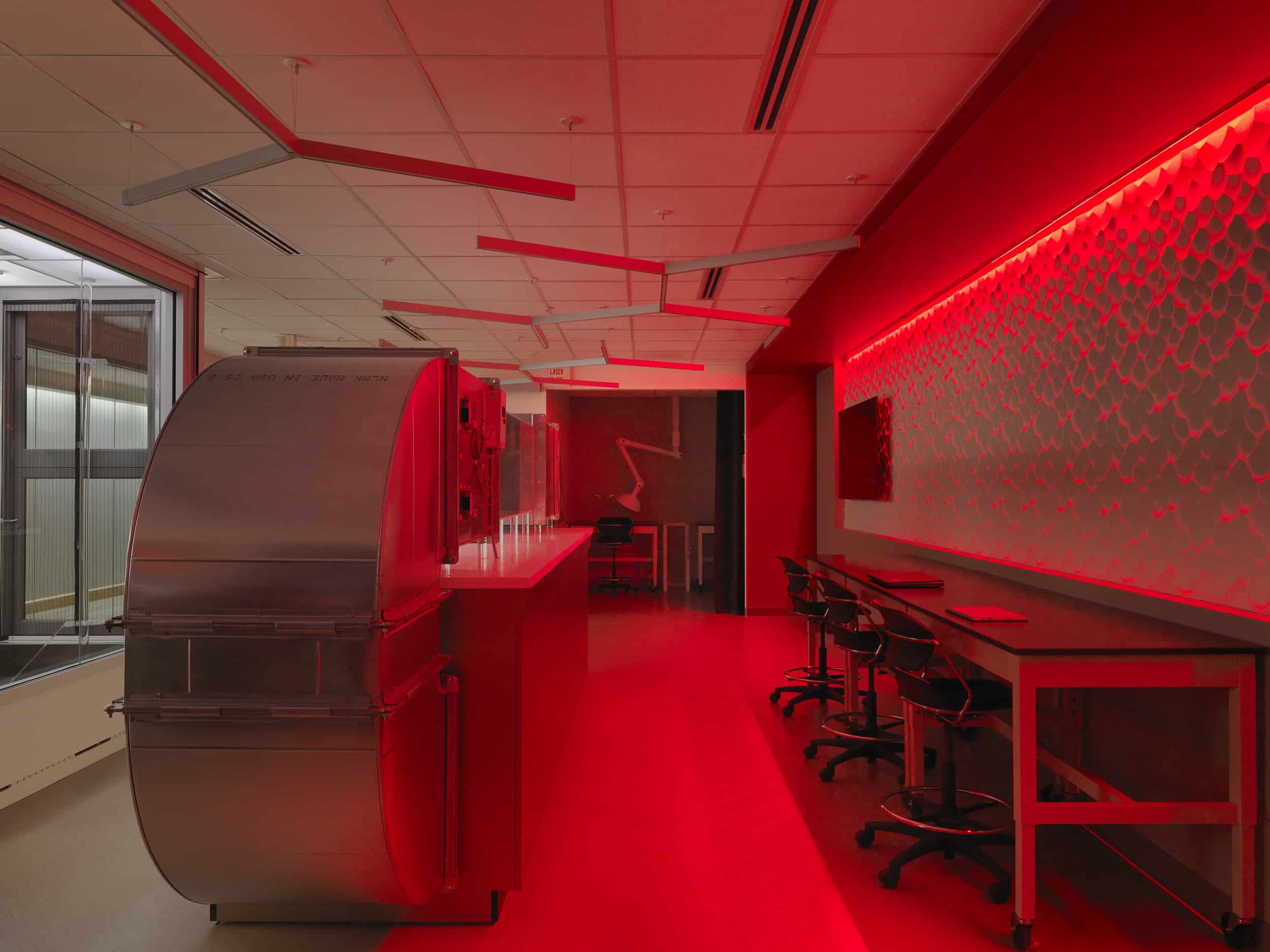 This image shows a laboratory space bathed in dramatic red lighting. The room features a white drop ceiling with geometric suspended panels, workstations with chairs along a counter on the right, and what appears to be specialized scientific equipment on the left side. The red illumination creates a striking, almost otherworldly atmosphere throughout the space. A textured wall panel or display is visible on the right wall, and large windows or glass partitions can be seen on the left side of the room.
