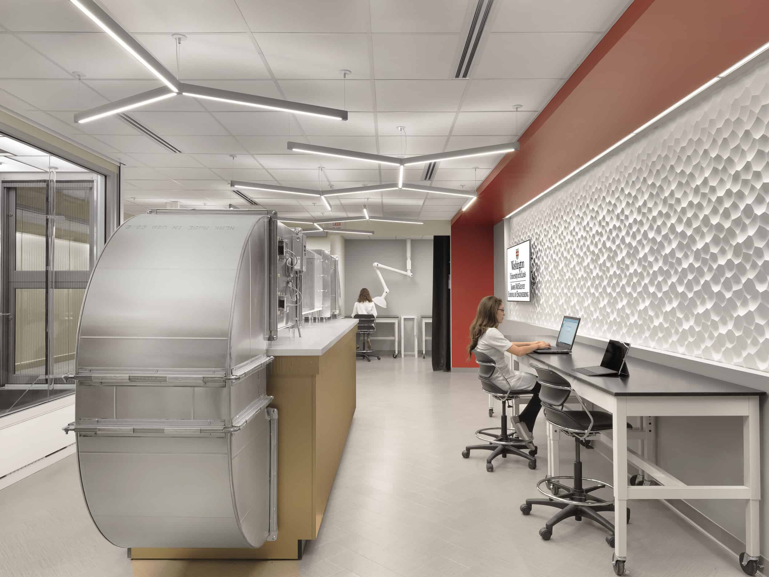 This image shows another view of the same laboratory space under normal lighting. The same cylindrical scientific equipment is visible on the left side, mounted on a wooden cabinet base. The textured hexagonal wall pattern with the red accent wall is visible on the right. Two individuals are working in the lab: one person is seated at a workstation along the right wall using a laptop computer, while another person can be seen in the background near what appears to be an articulated mechanical arm or instrument. The lab features the same geometric lighting fixtures on the ceiling and glass partitions on the left side. The modern workspace has a clean, professional appearance with plenty of counter space for research activities.