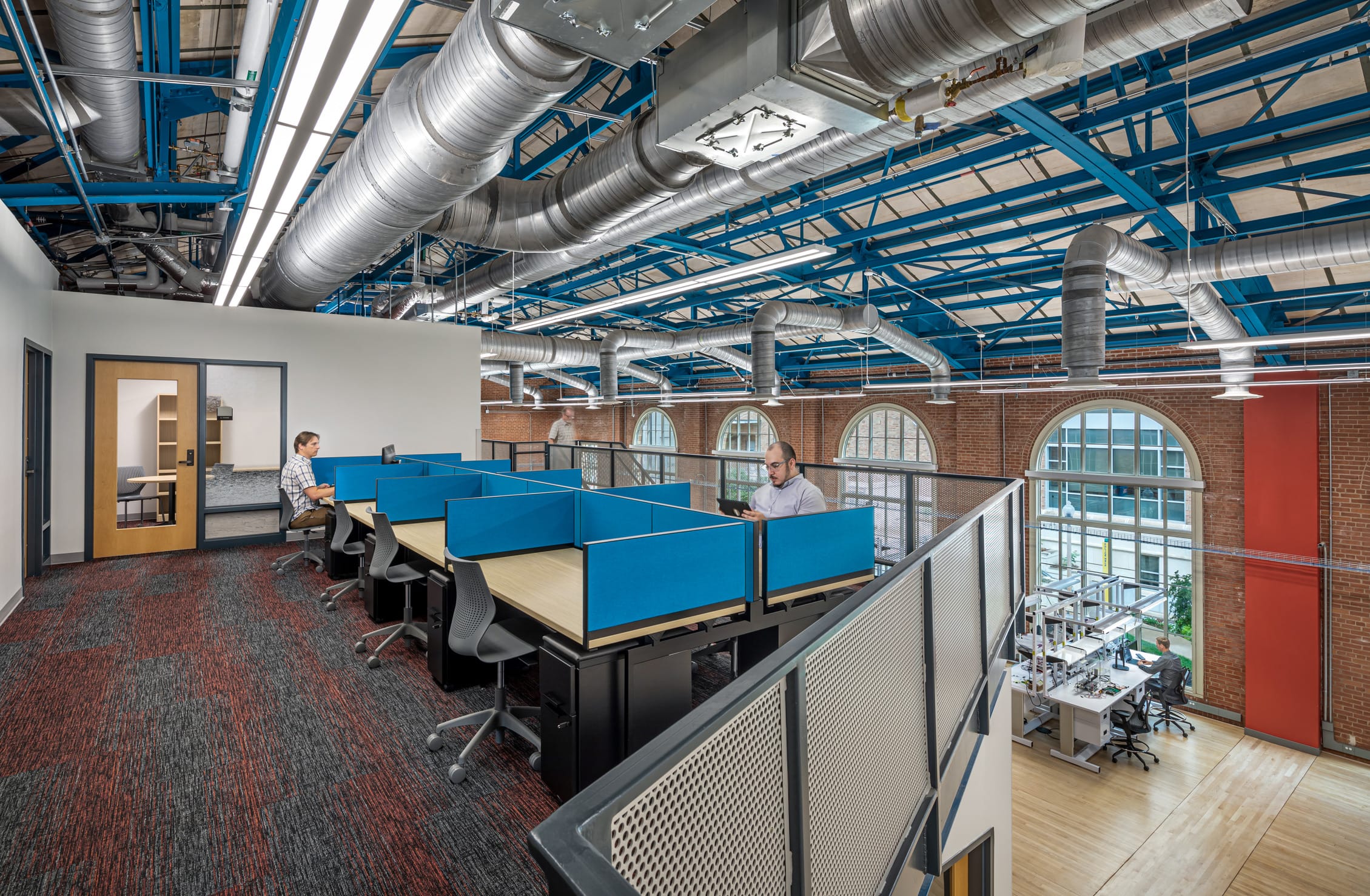 This image shows an industrial office space within the Anna Hiss Gymnasium-turned Robotics Lab with modern workspace elements. The interior features an open ceiling with exposed silver ductwork and blue metal framing, brick walls with large arched windows, and a multi-level workspace layout. On the upper level, there are modular workstations with blue dividers where two people are working at desks. Below is an open collaborative area with additional workstations. The space combines industrial elements with contemporary office design, featuring a mix of flooring types including carpeted and wooden areas.