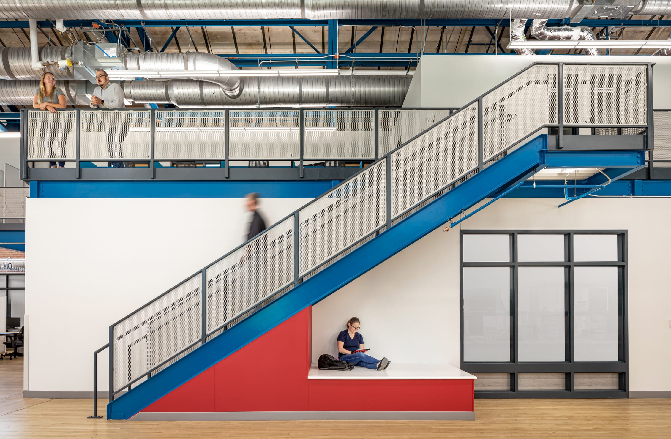 This image features a prominent blue staircase with perforated metal railings connecting two levels. Two people are conversing at the railing on the upper level beneath exposed metal ductwork and blue structural supports. On the lower level, a person in dark clothing is sitting on a built-in red and white bench/platform beneath the staircase, looking at a tablet or device. Another individual can be seen in motion on the stairs (appearing slightly blurred). The space maintains an industrial aesthetic with exposed ceiling systems, wooden flooring, and a minimalist design approach that combines functional elements with architectural interest. The combination of white walls, blue structural elements, and red accents creates a cohesive design language throughout the facility.