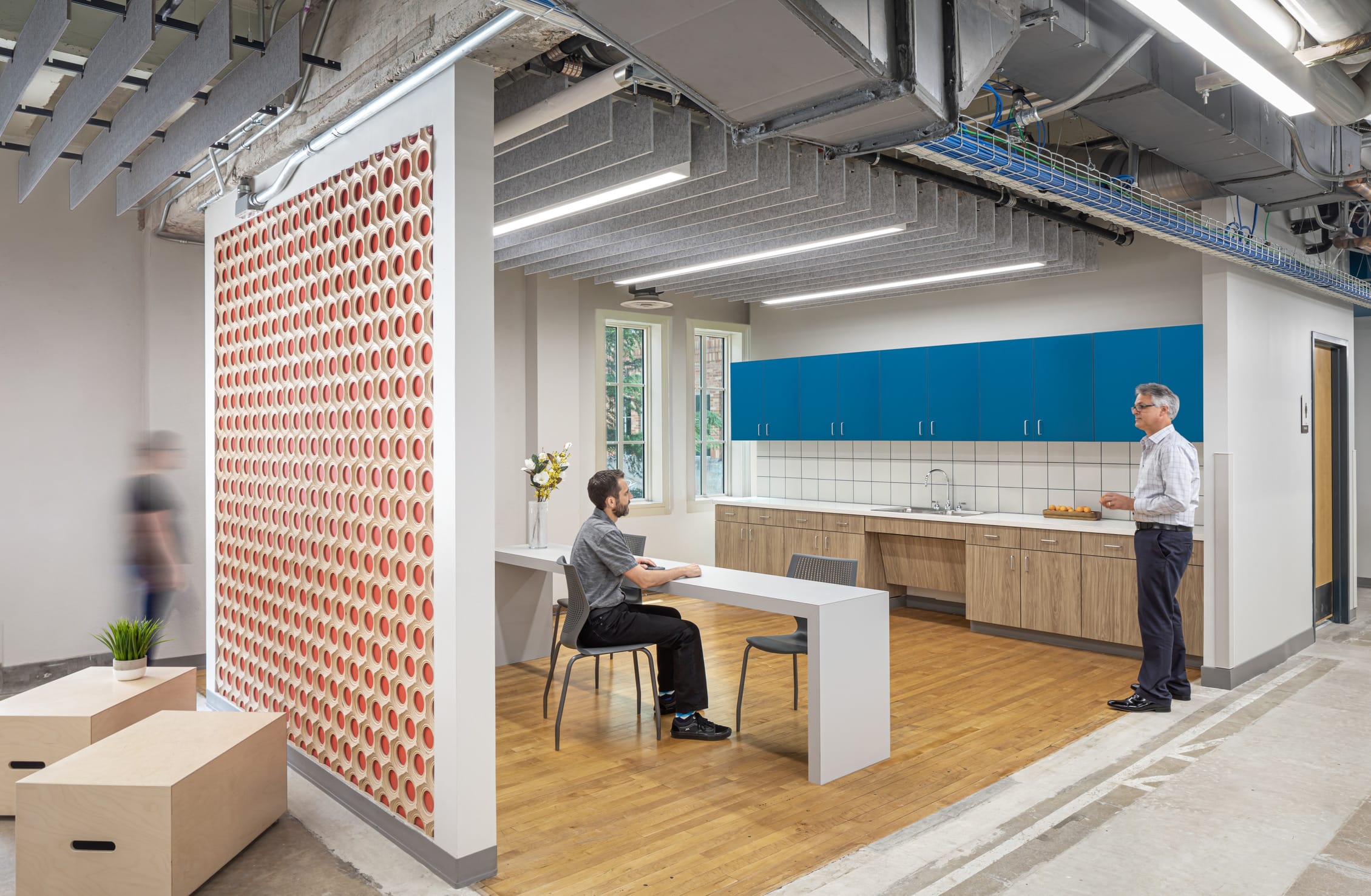 This image shows a break room or kitchen in modern workplace and educational facility. The space features bright blue upper cabinets and wooden lower cabinets with a white countertop and sink. A person with short dark hair sits at a simple white table in the center of the area, while another person with gray hair stands near the counter. The kitchen space is partially enclosed with a decorative red and white patterned wall panel on one side that adds visual interest. The ceiling maintains the industrial exposed look with visible ductwork, cables, and lighting fixtures. Natural light enters through windows visible in the background. The wooden flooring continues the aesthetic seen in previous images, while some wooden box-like seating or storage elements appear in the foreground. A small plant adds a touch of greenery to the space. A blurred figure can be seen walking past in the corridor.