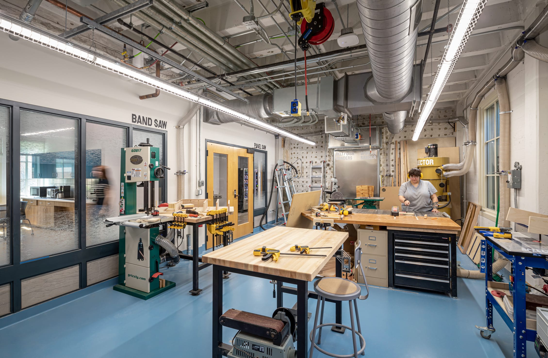 This image shows a well-equipped woodworking shop. The workshop features a blue floor and multiple woodworking stations with butcher block work surfaces. A green Grizzly band saw is prominently labeled on the left side of the room, with other workshop areas and equipment visible throughout the space. A person in a light gray shirt is working at one of the wooden workbenches on the right side. The space has industrial ceiling with exposed ductwork, ventilation systems, and electrical conduits. There's a retractable power cord reel visible hanging from the ceiling. Various power tools, clamps, and woodworking supplies are organized throughout the shop. Yellow doors with windows lead to other areas, and there's a glass partition on the left that looks into an adjacent space. The workshop appears to be designed for fabrication and prototyping work.