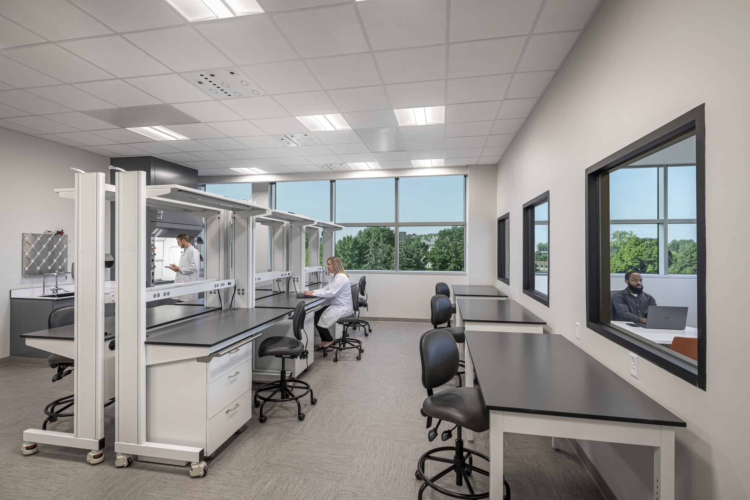 The image shows a modern laboratory or scientific workspace with multiple workstations arranged throughout a spacious room. Each station features black countertops mounted on white metal frames with adjustable height capabilities, complemented by black ergonomic chairs on wheels. The ceiling has a grid of fluorescent lighting panels and utility connections hanging down at various points. The room has light blue accent walls, frosted glass partitions on the left side, and appears to be designed for research or technical work with electrical outlets and equipment mounting points visible at each workstation. The space has a clean, organized appearance with neutral flooring and bright overhead lighting.