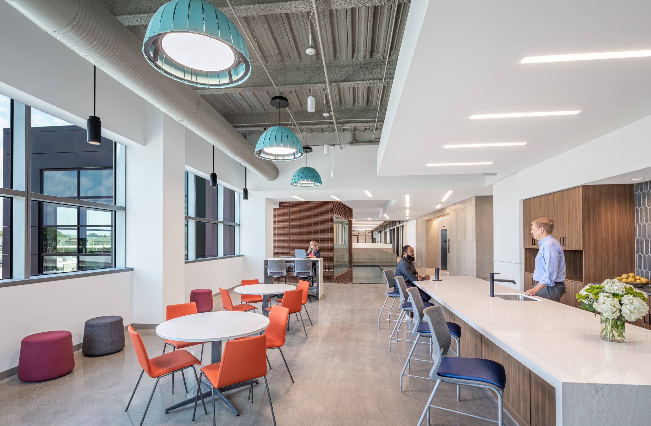 The image shows a modern office break room or café area with a bright, contemporary design. The space features white round tables with vibrant orange chairs in the foreground, alongside some burgundy and gray ottomans. To the right is a long countertop or bar with bar stools where one person with a beard is seated and another person in a blue shirt is standing. The counter area includes a sink, some flowers in a vase, and a bowl of fruit. The ceiling has an industrial feel with exposed ductwork and stylish turquoise pendant lights hanging down. Large windows along the left wall allow natural light to flood the space and provide views to the outside. In the background, there are additional seating areas including what appears to be a small meeting space with a woman sitting at a table, and some wooden slat partitions that create visual separation. The overall aesthetic combines wood elements, white surfaces, and colorful furniture to create an inviting collaborative space for employees.