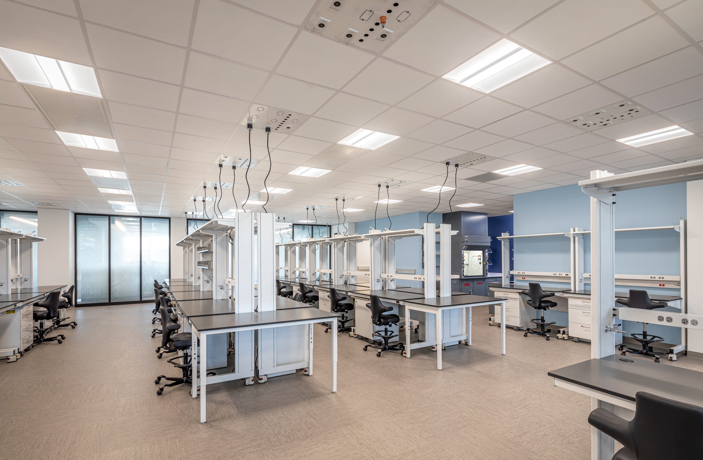 The image shows a modern laboratory or scientific workspace with multiple workstations arranged throughout a spacious room. Each station features black countertops mounted on white metal frames with adjustable height capabilities, complemented by black ergonomic chairs on wheels. The ceiling has a grid of fluorescent lighting panels and utility connections hanging down at various points. The room has light blue accent walls, frosted glass partitions on the left side, and appears to be designed for research or technical work with electrical outlets and equipment mounting points visible at each workstation. The space has a clean, organized appearance with neutral flooring and bright overhead lighting.