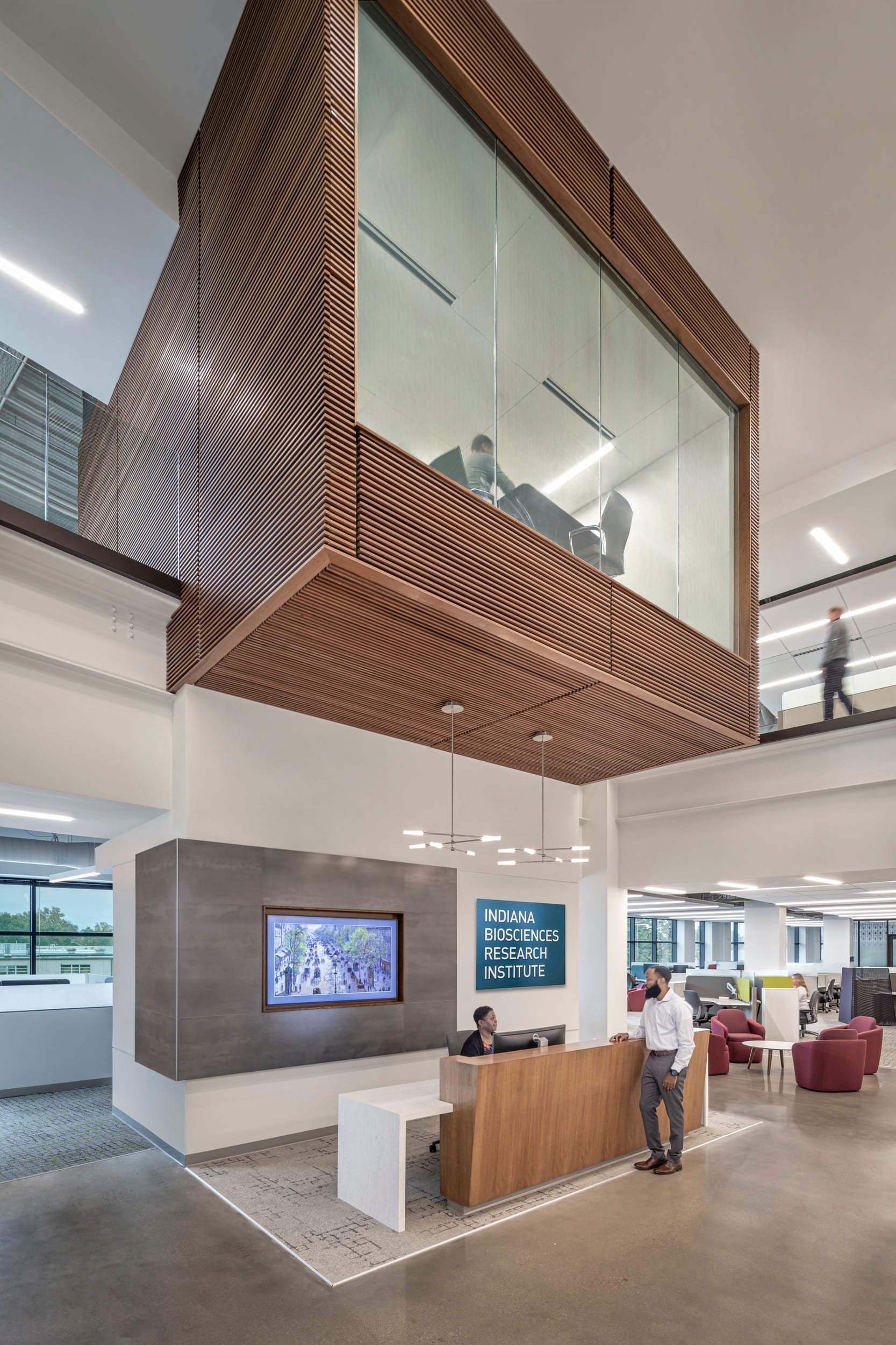 The image shows the reception area of the Indiana Biosciences Research Institute. The architectural design features a striking wooden cantilevered meeting room or conference space suspended above the reception desk. This floating box structure is clad in warm wooden slats with large glass panels that allow visibility into the meeting space where some people can be seen. Below, there's a modern reception desk with a wooden front where a person is sitting, and a man in business attire is standing at the counter. The reception area includes a dark gray accent wall with a built-in digital display screen. A blue sign with