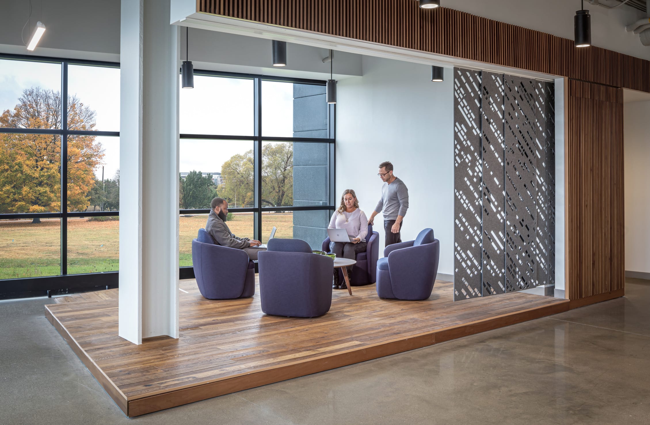 The image shows a modern open office environment with an interesting architectural design that balances privacy and openness. On the left side, there's a wall of frosted glass panels with a pattern of small dots, bordered by wooden panels. The ceiling features exposed pipes, conduits, and ductwork in some areas, with drop ceiling panels in others, creating a contemporary industrial aesthetic. The office space contains cubicle-style workstations with low dividers. Two people are visible working at their desks - a woman on the right side seated at her workstation and a person working at a desk with a bright yellow privacy divider behind them. A person in a blue shirt is walking through the hallway on the left side of the office. The floor has patterned carpeting with a subtle geometric design in beige and brown tones. The space appears bright and airy with natural light coming through windows visible in the background, and the overall design balances modern office functionality with aesthetic elements.Three people are working in the space - a bearded man sitting in a purple chair with a laptop, a woman with blonde hair also working on a laptop, and a man in glasses standing nearby. They're arranged around comfortable purple lounge chairs and a small round table in a casual meeting setup. The area has floor-to-ceiling windows along one wall providing views of autumn trees with orange and yellow foliage outside. On the right side, there's a decorative metal screen with a geometric pattern that provides partial privacy while maintaining an open feel. The ceiling features wooden slat details and pendant lighting fixtures. The overall design balances openness with privacy, creating an inviting space for informal meetings or collaborative work.