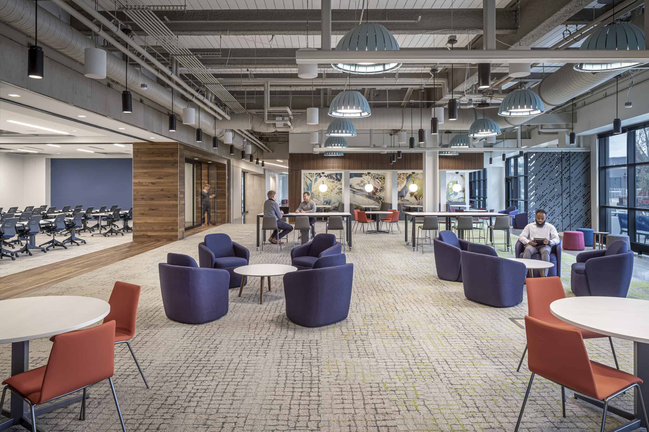 The image shows a modern collaborative workspace with multiple sets of tables and chairs in different configurations. One person is in comfortable purple lounge chairs and a small round table in a casual meeting setup. Two other men are sitting at a high-top table on grey chairs and collaborating over a laptop. The overall design balances professional and casual elements with a mix of materials including wood, concrete, and fabric, creating a versatile workspace that supports different types of interactions and activities.