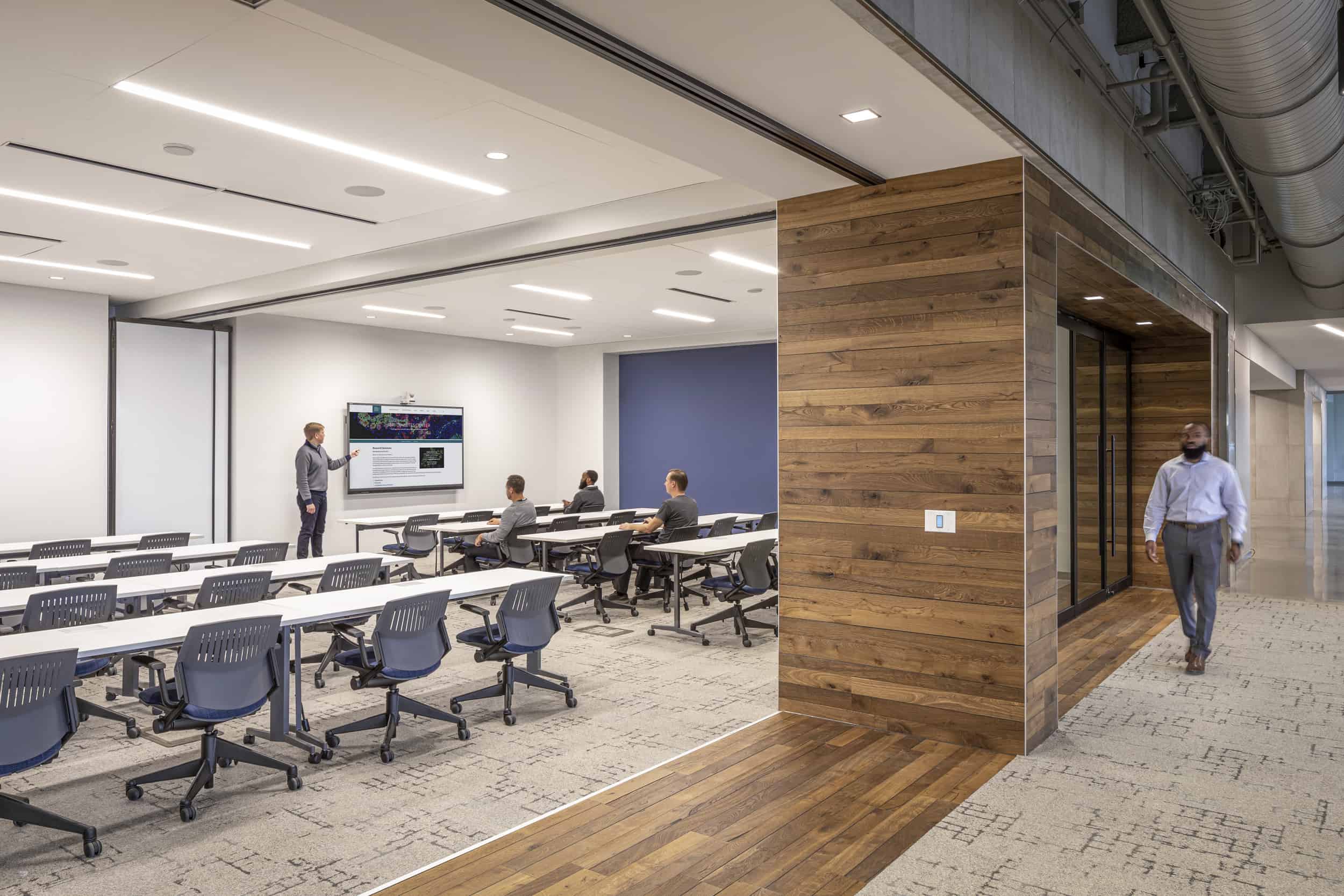 The image shows a modern open office environment with an interesting architectural design that balances privacy and openness. This view is of a meeting/presentation room with eight tables arranged neatly in two rows of four. Three people are sitting at the tables, which each have four ergonomic chairs, and watching a man deliver a presentation on a large flat screen TV. The entrance to the room is a dramatic wooden arch, but the walls are transparent glass. The floor has patterned carpeting with a subtle geometric design in beige and brown tones.