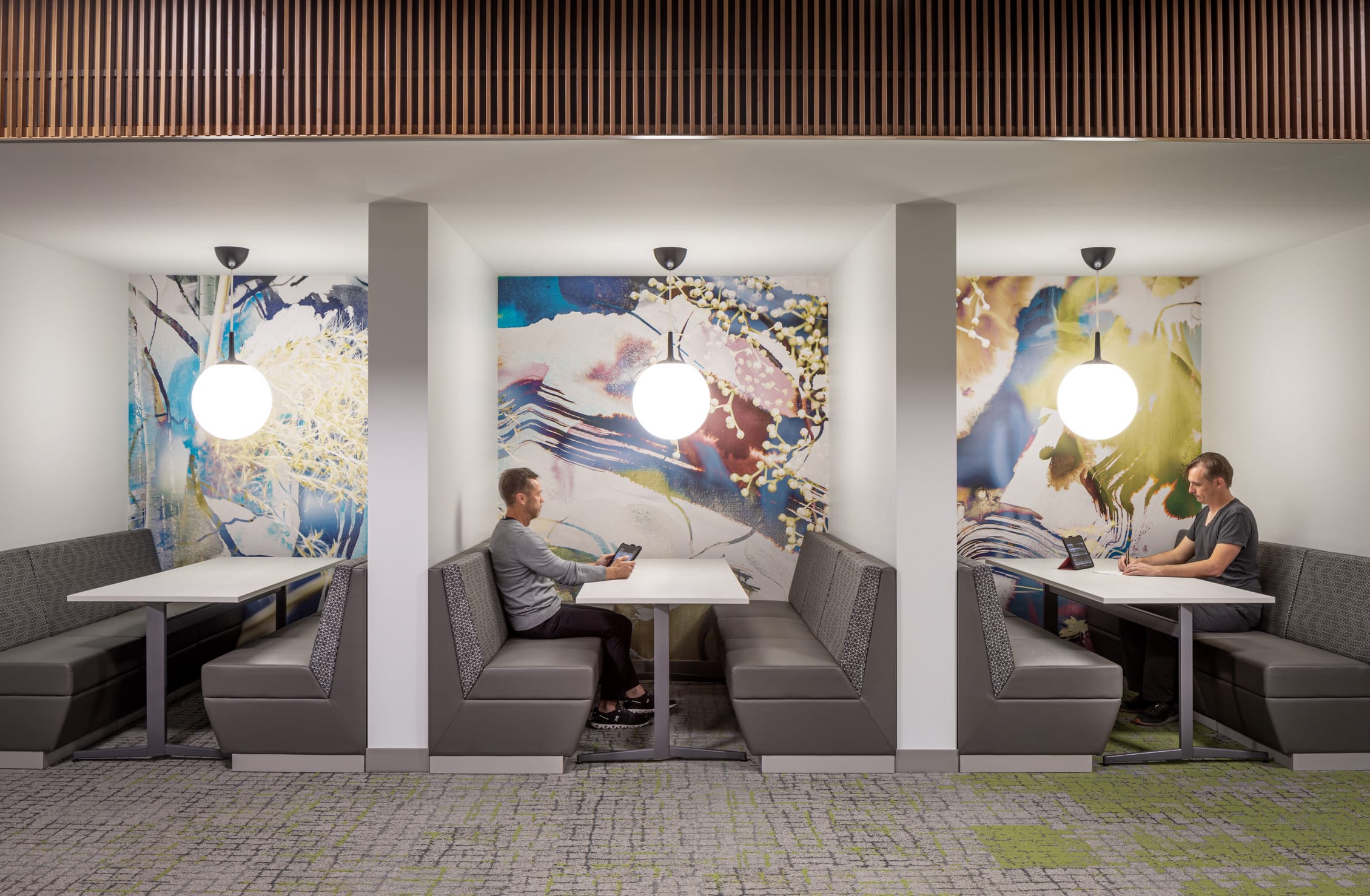 The image shows a modern office space with three side-by-side booth seating areas designed for focused individual work or small meetings. Each booth features gray upholstered bench seating arranged in a U-shape around white tables. Two of the booths are occupied by men working on tablets or laptops. What makes these booths visually striking is the colorful abstract artwork that covers the back wall of each alcove - featuring flowing, organic designs in blues, yellows, and multicolored tones. Each booth is illuminated by a single spherical pendant light hanging from the ceiling. The top of the image shows wooden slat detailing along the ceiling, and the floor features a geometric patterned carpet in gray with some lime green accents. The overall design creates semi-private workspaces that blend functionality with artistic elements, allowing for concentration while maintaining an open feel to the environment.