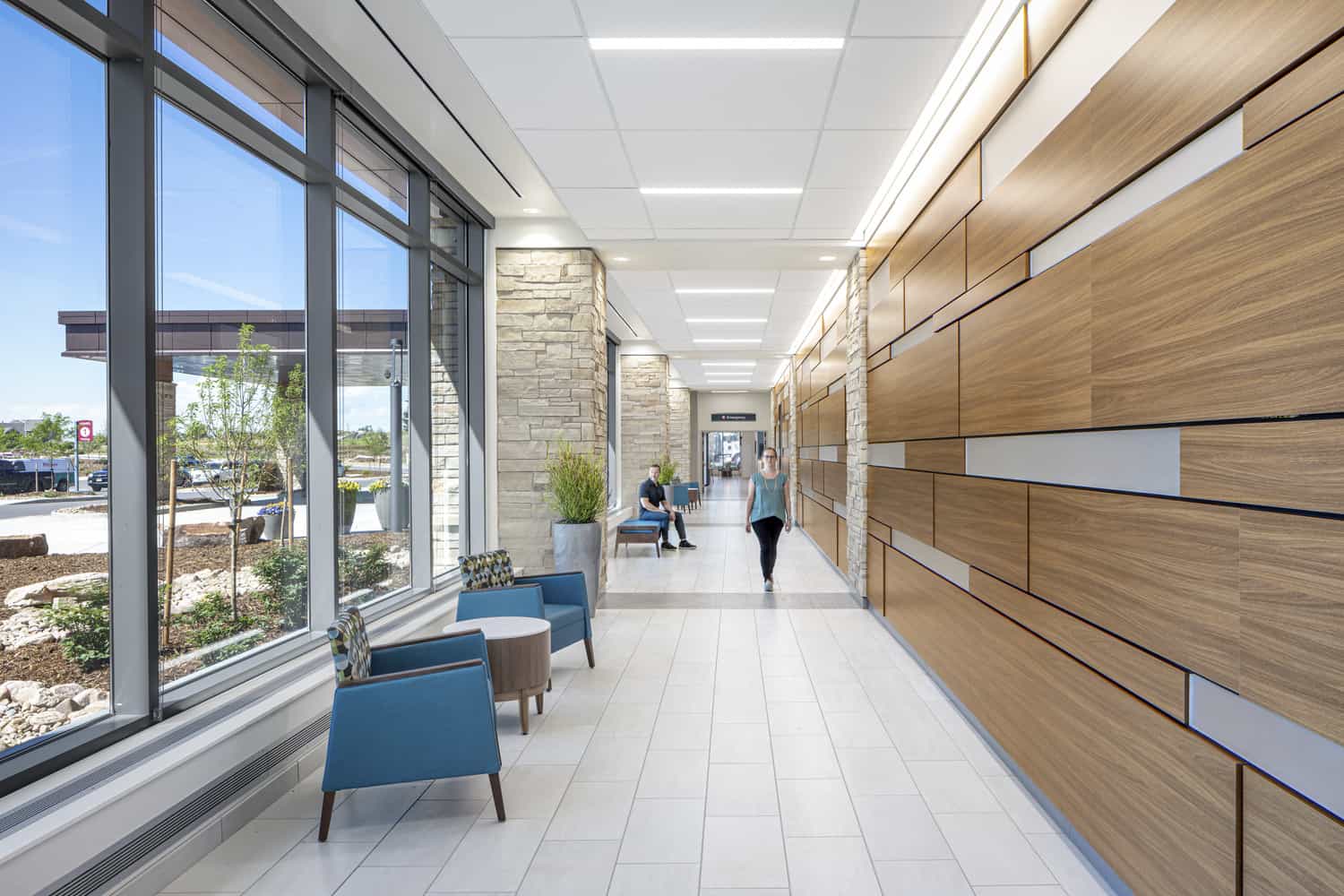 Modern hallway within the UCHealth Greeley Hospital. The space features floor-to-ceiling windows overlooking a parking lot and landscaped grounds. Blue armchairs and a round wooden table are positioned near the windows. The opposite wall is made of attractive wood panels protruding at different levels, creating a textured design.