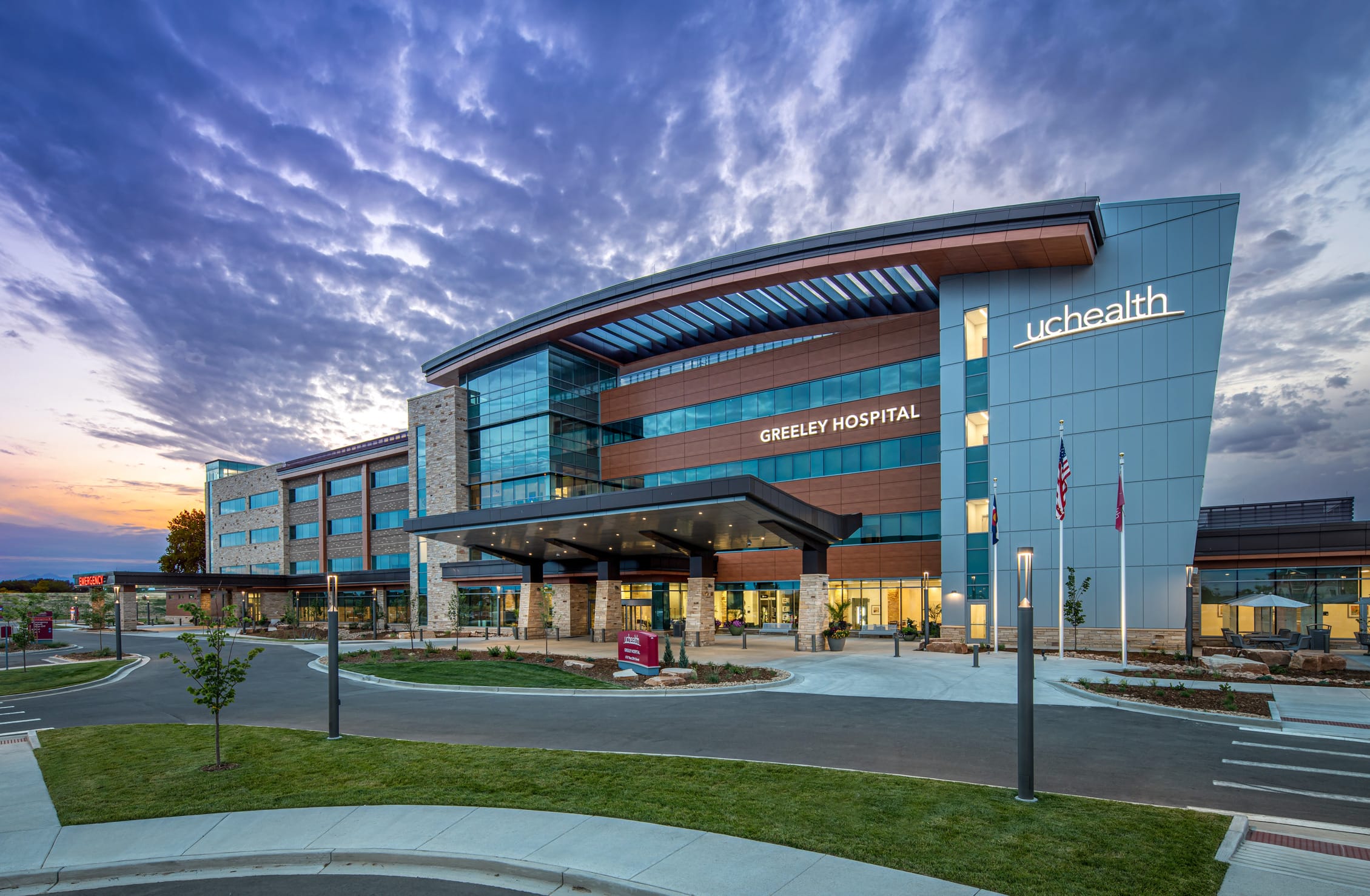 A modern hospital building labeled 'Greeley Hospital' with the 'UCHealth' logo, captured at dusk. The architecture features a combination of glass, metal, and stone materials, with a curved roof and large windows reflecting the sky. The entrance has a well-lit covered drop-off area supported by stone pillars. Three flagpoles stand in front of the building, displaying the American flag and other flags. The surrounding landscape includes neatly trimmed grass, small trees, and decorative lighting. This image shows BSA's expertise in healthcare architecture.