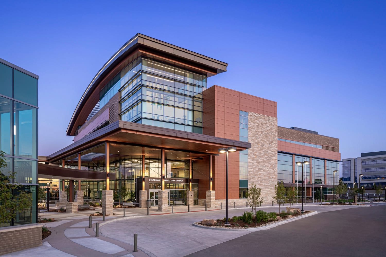 This image shows the entrance to UCHealth's Steadman Hawkins Clinic, which is a modern, multi-story building with a curved roof and extensive glass windows. The entrance architecture combines a curved glass atrium, brick, and wood elements, with a large wooden canopy. This image shows BSA's expertise in striking and unique architecture.