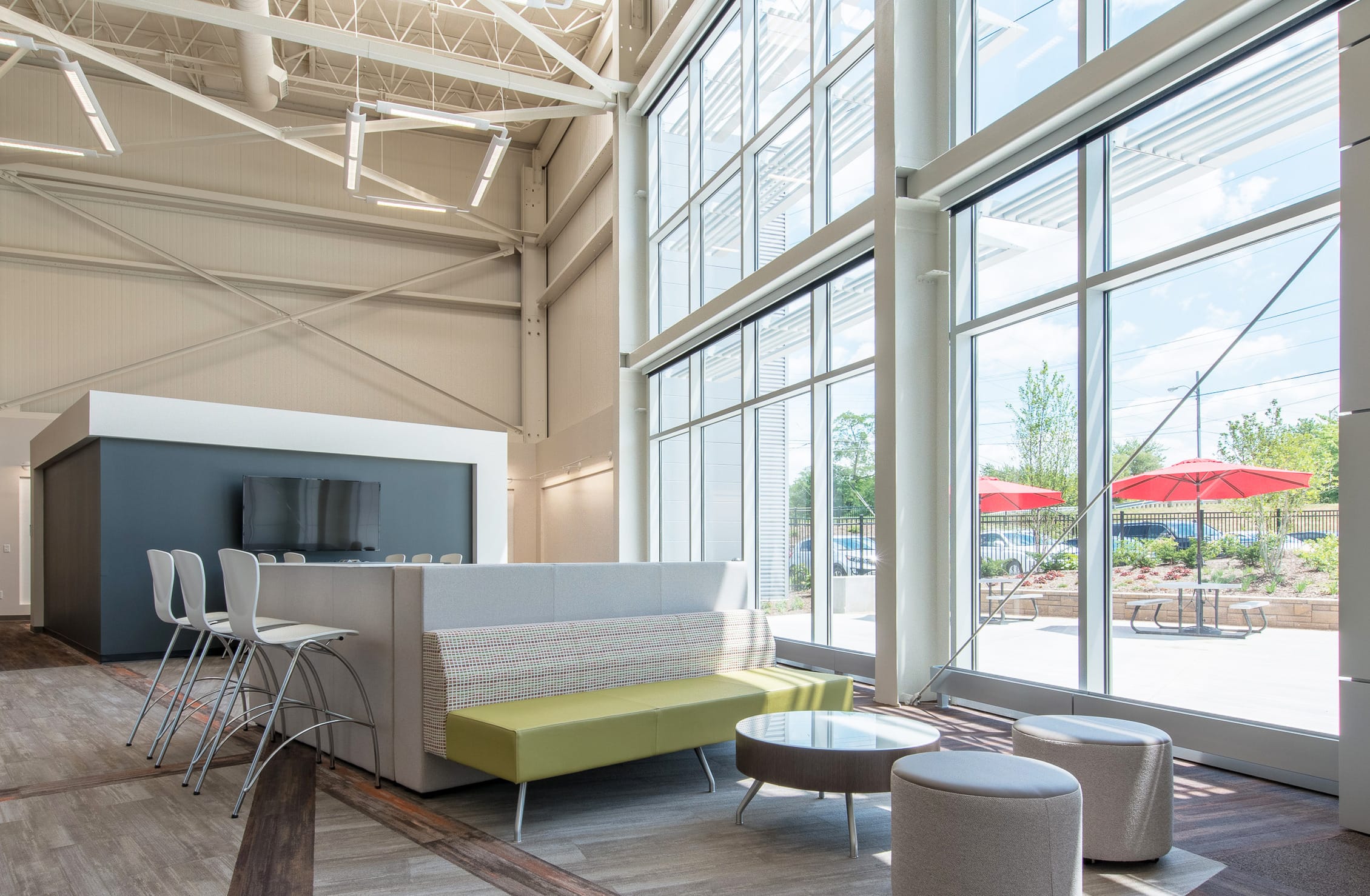 A spacious, modern office break area with high ceilings and floor-to-ceiling windows. Outside, red umbrellas and greenery are visible through the expansive glass windows, adding to the bright, airy atmosphere. There are multiple pieces of modern furniture in lime green and grey arranged in different configurations. Overhead, modern lighting and exposed ductwork contribute to the contemporary, open-office aesthetic.