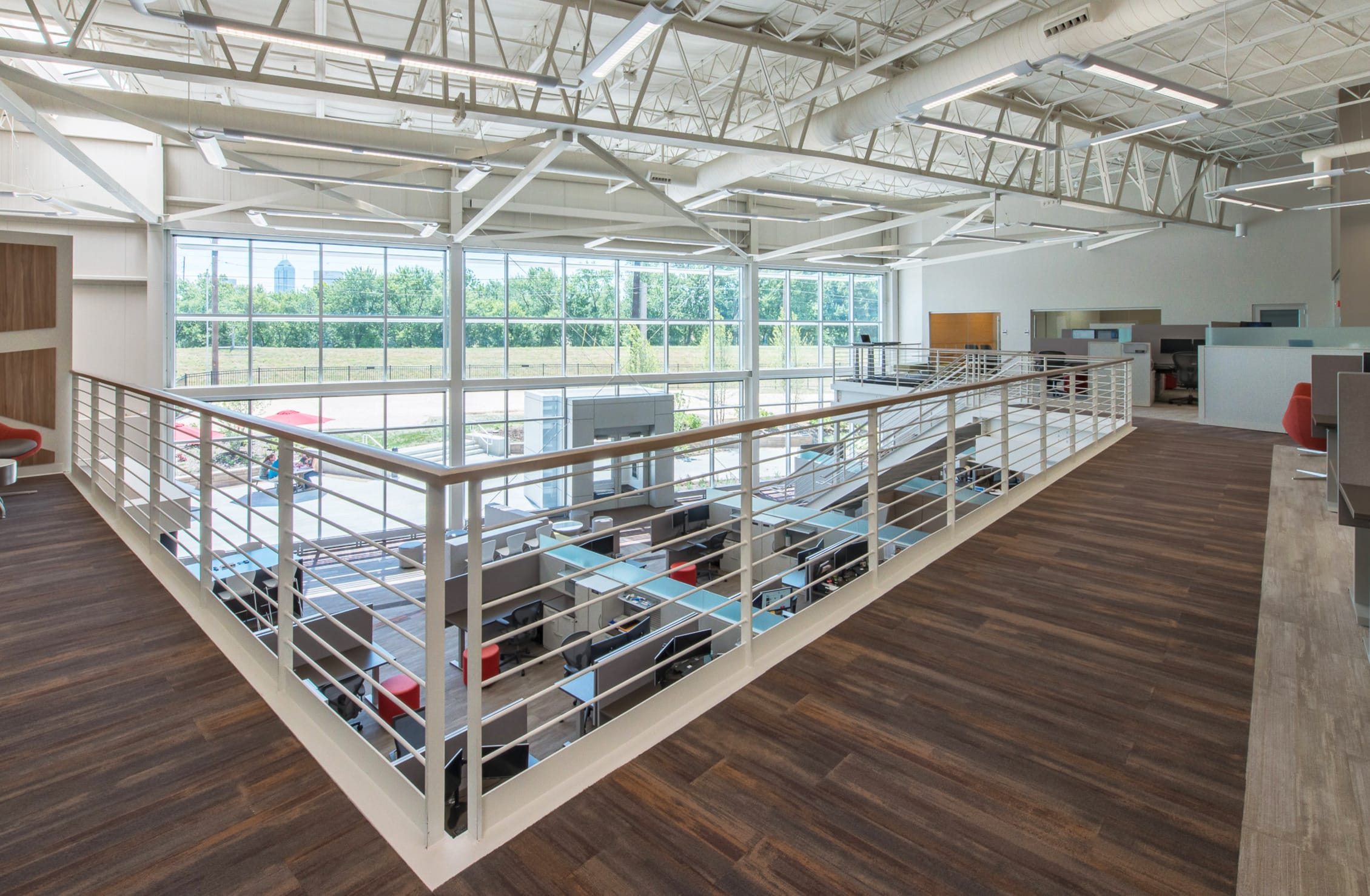 Interior of a modern, open-concept office building with a mezzanine level overlooking a workspace below. The space features high ceilings with exposed white steel trusses and large floor-to-ceiling windows that let in abundant natural light. The mezzanine has wood-look flooring and a white metal railing. Below, cubicles with glass dividers and black office chairs are arranged neatly. Outside, red umbrellas and greenery are visible through the expansive glass windows, adding to the bright, airy atmosphere.