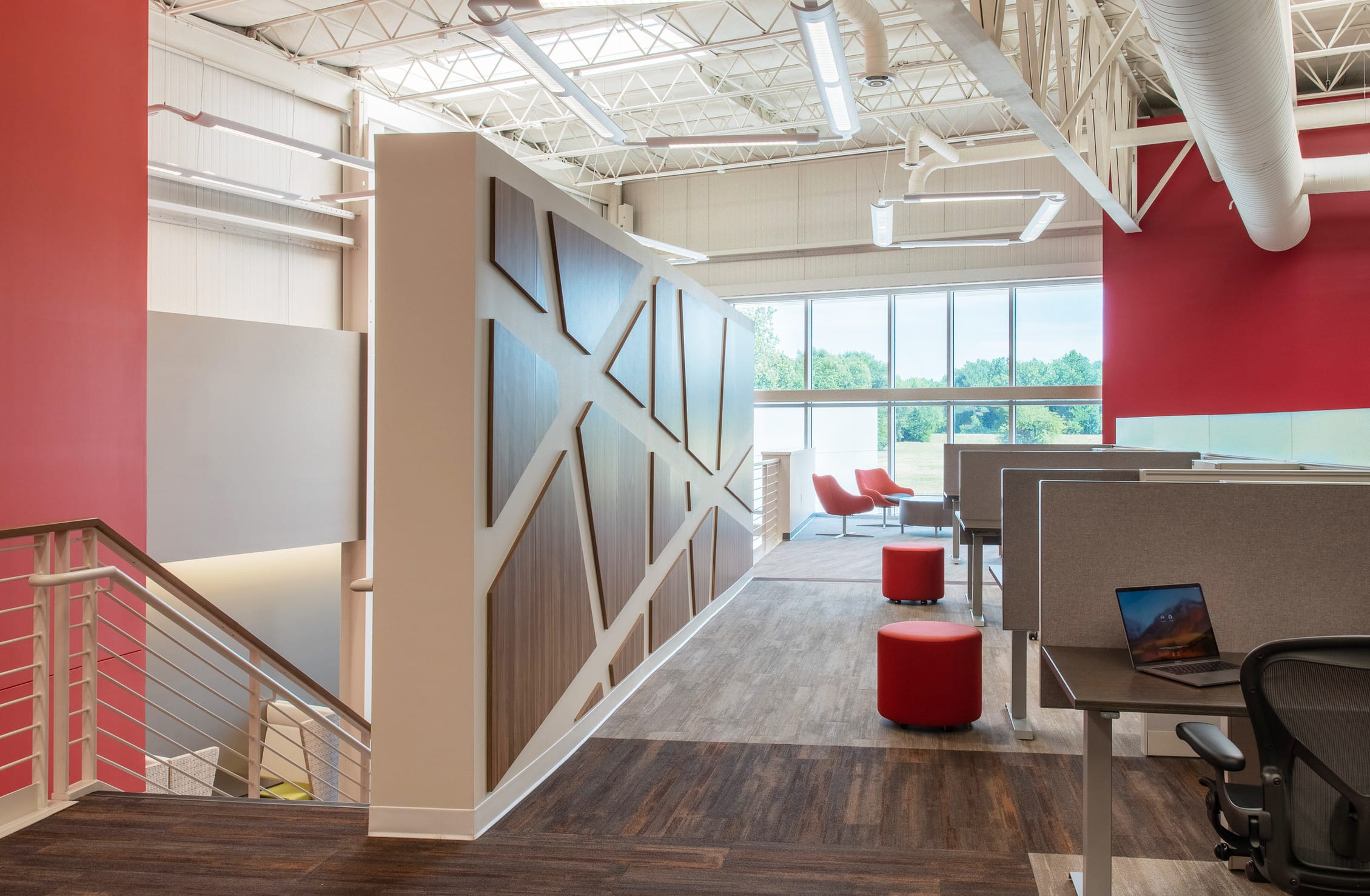 A spacious, modern upstairs office work area with cubicles and a warm, welcoming color palette. The space features a central staircase with geometric wooden paneling on the wall beside it. The cubicles offer some privacy with fabric grey dividers, and some modern red chairs and ottomans are positioned randomly outside of workspaces. Overhead, modern lighting and exposed ductwork contribute to the contemporary, open-office aesthetic.