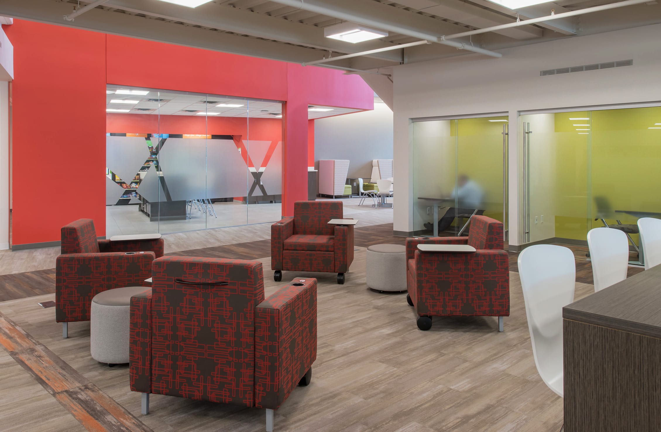 A spacious, modern office break area with high ceilings and a bright, welcoming color palette. The space features four central chairs with a tech-like pattern on them neatly arranged. To the right are two private meeting rooms with frosted glass doors for some privacy. These rooms stand out amongst the grey break area walls because they are bright green. Another break room with what looks to be vending machines is in the back, also standing out with bright red paint. Overhead, modern lighting and exposed ductwork contribute to the contemporary, open-office aesthetic.