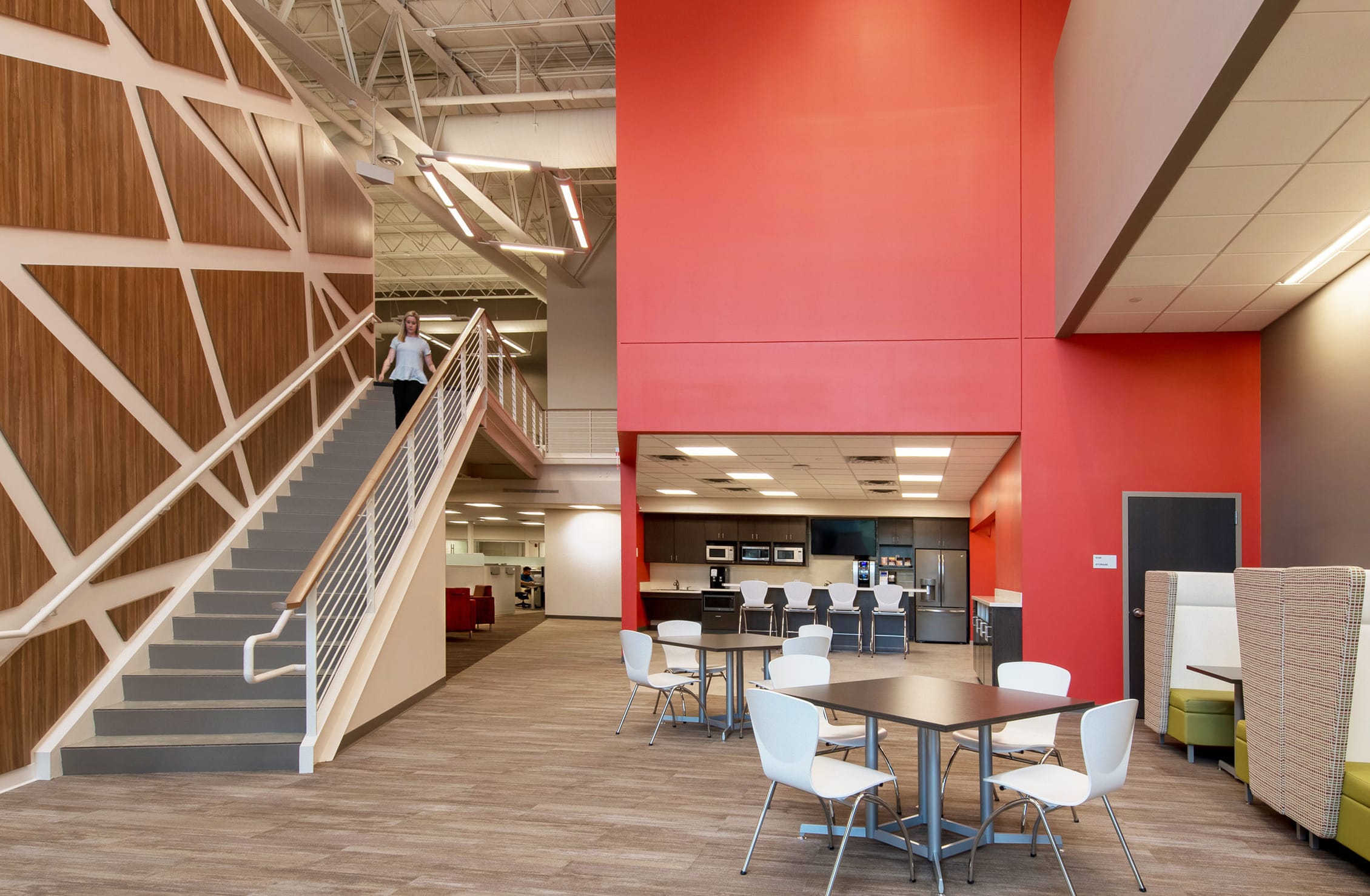 A spacious, modern office break area with high ceilings and a warm, welcoming color palette. The space features a central staircase with geometric wooden paneling on the wall beside it. A woman is descending the stairs. To the right, there are café-style tables and chairs, along with semi-enclosed booth seating upholstered in neutral tones. In the background, a large kitchen area includes multiple microwaves, a coffee machine, a water dispenser, and a fridge, all set against a red accent wall. Overhead, modern lighting and exposed ductwork contribute to the contemporary, open-office aesthetic.
