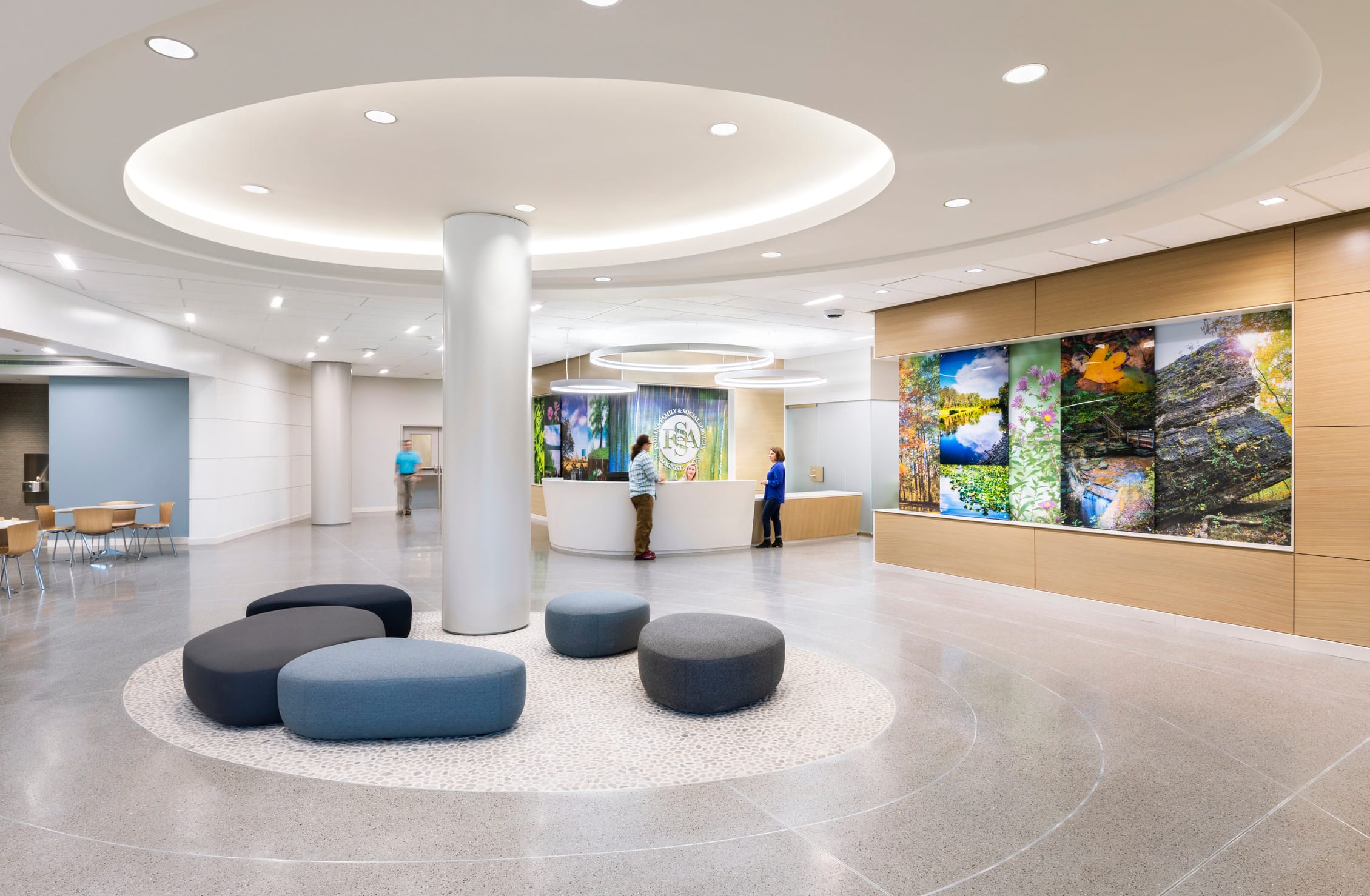 A modern and spacious lobby with a curved ceiling design featuring recessed lighting. The floor is polished with circular patterns complementing the seating area, which consists of round and pebble-shaped cushioned seats in shades of blue and gray. A white reception desk with a curved front is staffed, and a large illuminated logo behind it reads 'FSA.' The walls feature warm wood paneling and a large display of vibrant nature-themed artwork. In the background, a seating area with wooden chairs and tables is visible, along with a hallway leading further into the facility. The interior highlights BSA's expertise in environments that prioritize healing and patient well-being.
