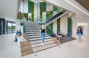 Modern interior of a Wake Tech Community College building featuring a bright, open lobby with a prominent staircase. The space has white floors and walls accented with green panels and living moss wall installations. A central staircase with wooden steps and glass railings connects floors, with several students in motion on the stairs. To the left, two students sit on built-in seating near large windows overlooking greenery outside. To the right, a small seating area with blue chairs and small tables includes a student studying. The design emphasizes natural light, open space, and biophilic elements that bring nature indoors.