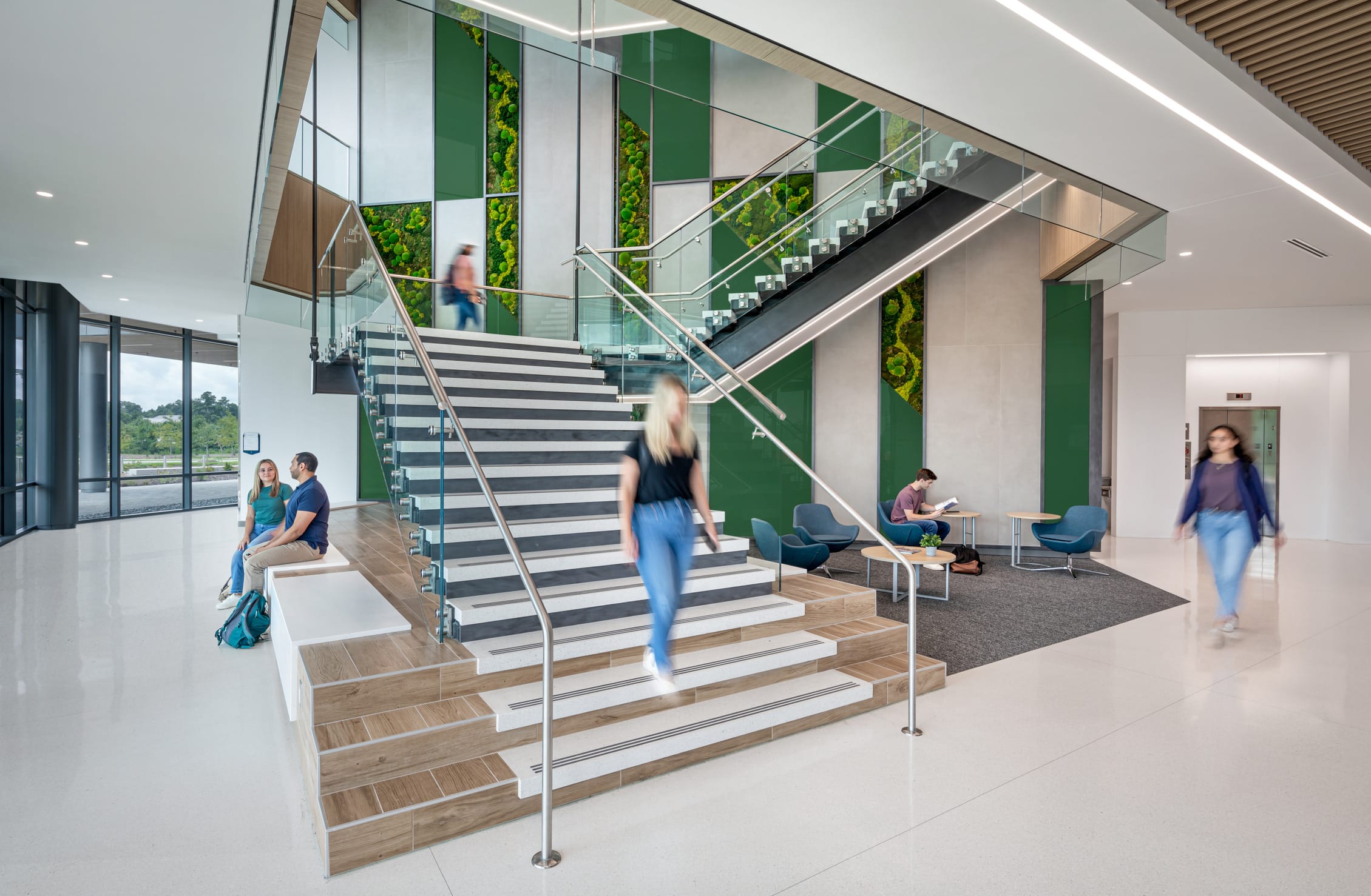 Modern interior of a Wake Tech Community College building featuring a bright, open lobby with a prominent staircase. The space has white floors and walls accented with green panels and living moss wall installations. A central staircase with wooden steps and glass railings connects floors, with several students in motion on the stairs. To the left, two students sit on built-in seating near large windows overlooking greenery outside. To the right, a small seating area with blue chairs and small tables includes a student studying. The design emphasizes natural light, open space, and biophilic elements that bring nature indoors.