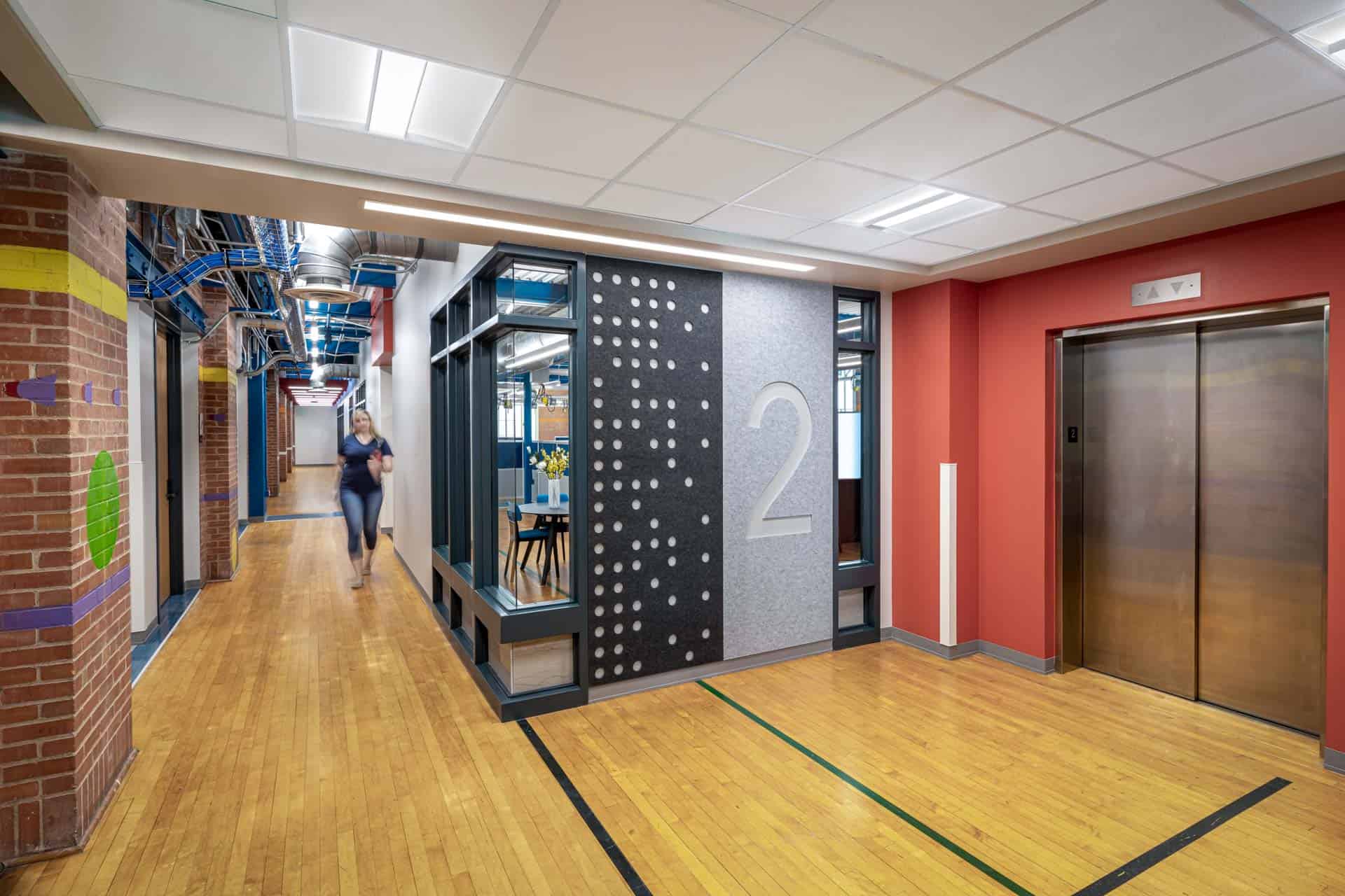 This image shows the now-modern interior of the renovated Anna Hiss Gymnasium with an elevator lobby and corridor intersection. The space features wooden flooring with directional lines inlaid, and a large number