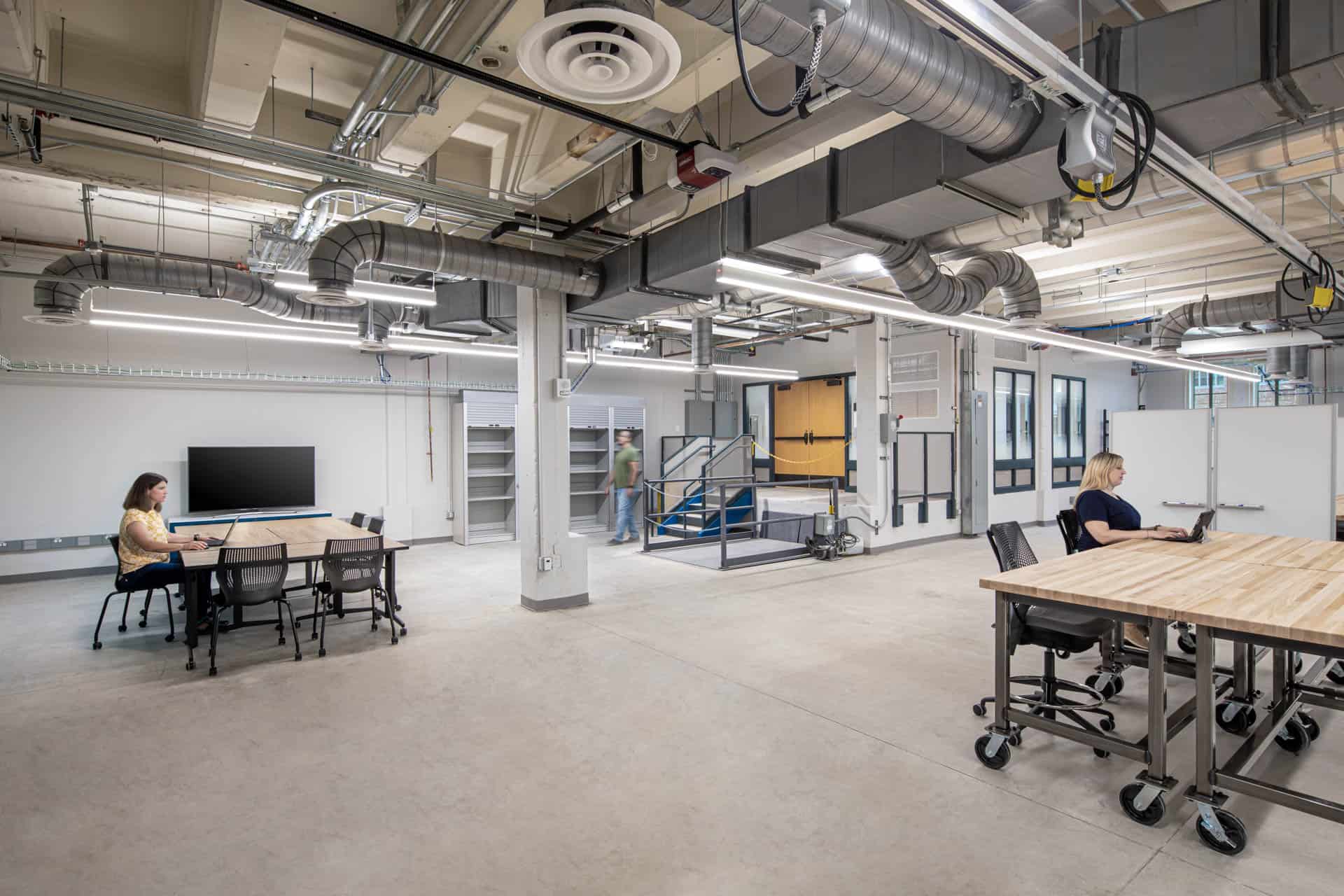 This image shows an industrial-style collaborative workspace with exposed ceiling infrastructure. The space features a polished concrete floor and white walls. Several people are visible - one woman sitting at a workstation on the left, one man entering from a door in the background, and one woman working on a computer at a workstation on the right. Near the back doorway is a large electric lift for large equipment. The ceiling reveals exposed ductwork, pipes, and lighting fixtures, creating an urban loft aesthetic. The minimalist design combines functional elements with modern workspace concepts, suggesting this may be part of a converted industrial building now serving as a creative or tech office environment.