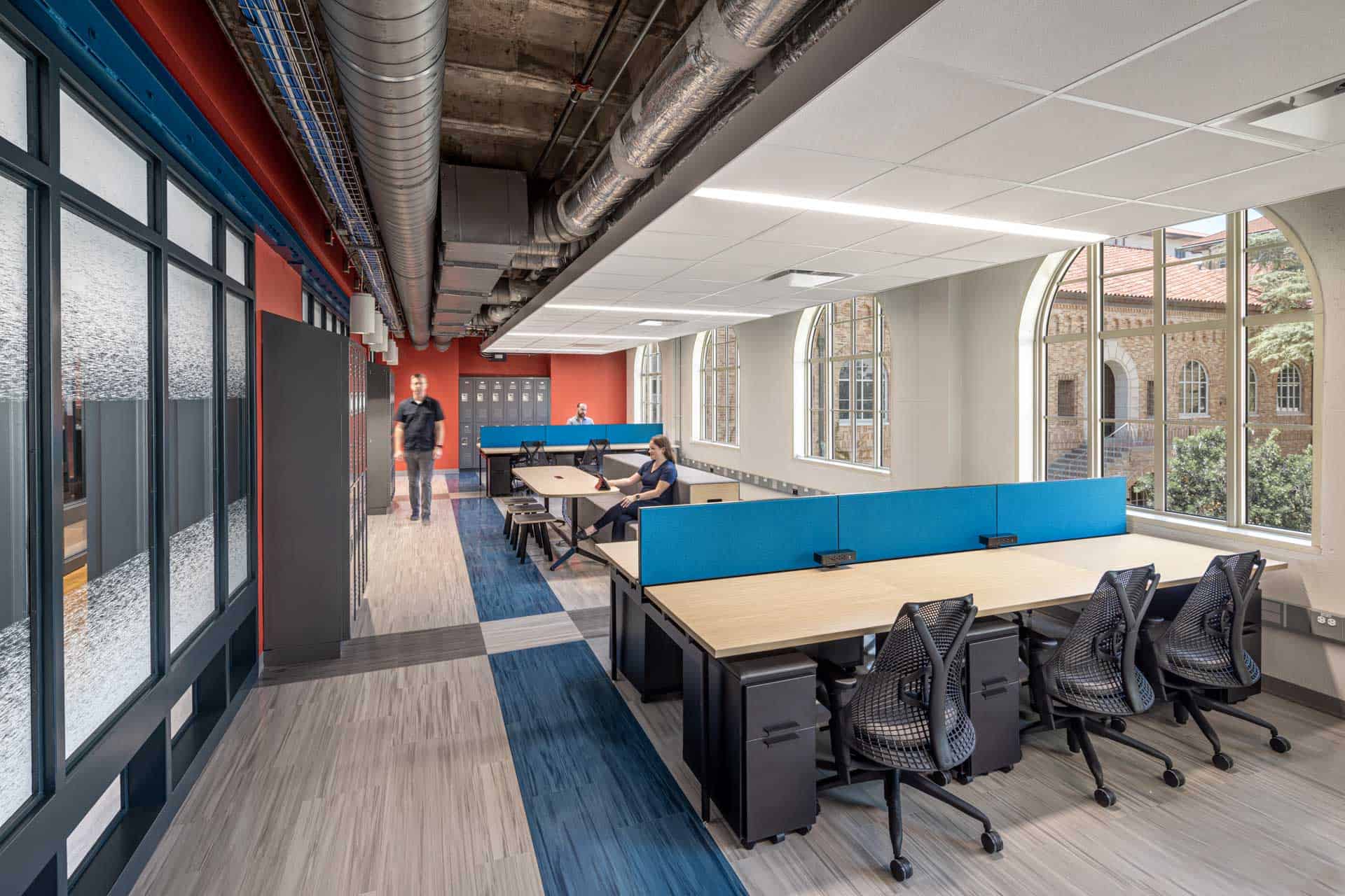 This image shows an industrial office space within the Anna Hiss Gymnasium-turned Robotics Lab with modern workspace elements. The interior features an open ceiling with exposed silver ductwork and large arched windows. There are modular workstations with blue dividers and a set of grey lockers, indicating this space is likely designed to be utilized by students.