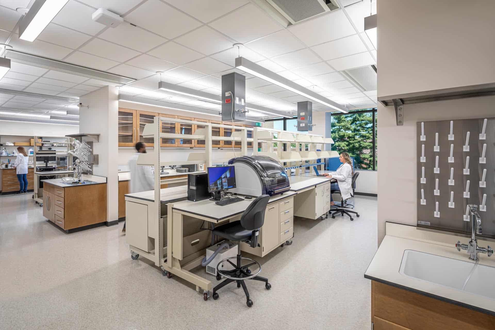 The image shows a well-equipped scientific laboratory with multiple workstations. The lab features wooden and beige cabinetry with white countertops organized in rows. Several analytical instruments are positioned on the counters, as sink and eye-wash stations. A computer workstation is visible with a monitor and keyboard, positioned at a high desk with a black rolling lab chair. Above the countertops are extensive open shelving and cabinets with glass doors allowing for visibility of materials. The space is well-lit with fluorescent ceiling lights, and a large window is visible on the back wall. The overall appearance is of a modern, functional research or testing laboratory in an academic or industrial setting.