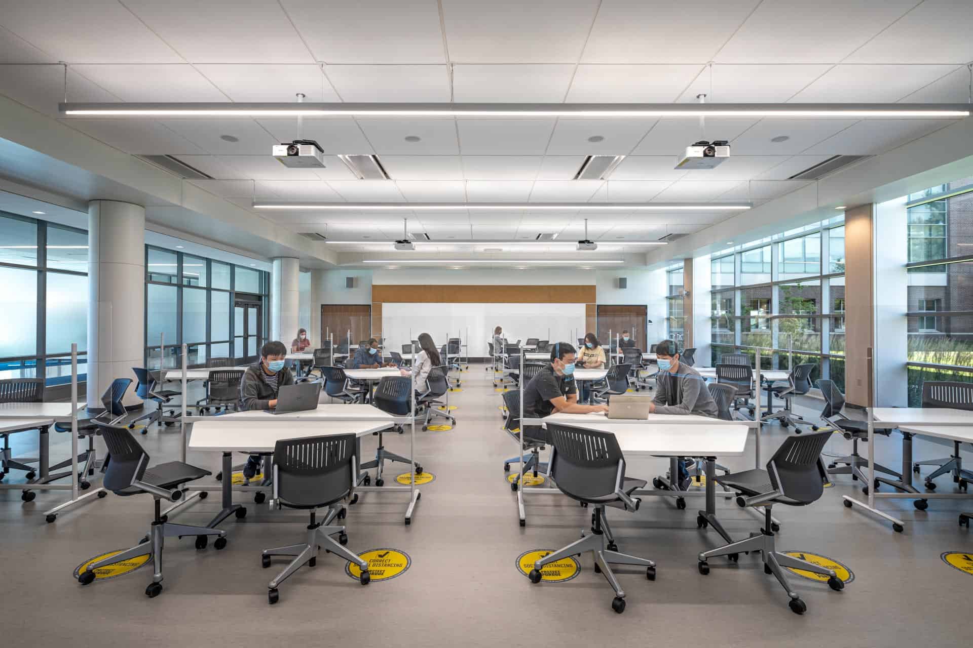 The image shows a modern educational or collaborative workspace during what appears to be the COVID-19 pandemic period. Several people wearing face masks are working at hexagonal or angular white tables that are spaced apart throughout a bright, open room. Yellow social distancing markers are visible on the floor around each workspace. The space has a contemporary design with large windows along both sides allowing natural light to enter, and ceiling-mounted projectors for presentations. The furniture includes black ergonomic chairs on wheels positioned at the white work tables. A few individuals are working on laptops throughout the room. In the background, there appears to be a presentation area or additional workspace with a light-colored wall. The overall impression is of a flexible learning environment or collaborative workspace that has been adapted for pandemic safety protocols, with intentional spacing between workstations and visual indicators for maintaining distance.