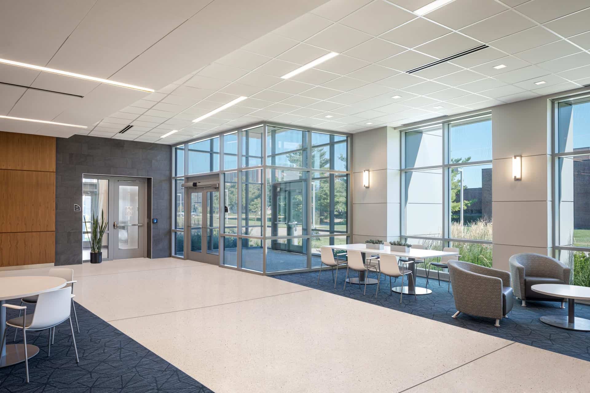 The image shows a modern interior lobby or entrance area of the Martin C. Jischke Hall. The space features a bright, open design with floor-to-ceiling glass walls and doors that serve as the main entrance, allowing abundant natural light to flood the interior. The lobby has a contemporary aesthetic with a mix of flooring materials - primarily light-colored terrazzo or polished concrete in the central area with patterned blue carpeting defining seating zones. Various seating arrangements are visible, including small round tables with white chairs on the left side and more comfortable lounge-style seating with upholstered gray chairs on the right. The walls feature a combination of materials including wooden paneling on the left and gray stone or tile accents. Wall-mounted sconce lighting provides additional illumination to complement the recessed ceiling lights. Through the glass walls, you can see landscaped grounds outside with some greenery and pathways. This appears to be a welcoming reception or gathering area designed to connect interior spaces with the outside environment while providing comfortable areas for brief meetings or waiting.