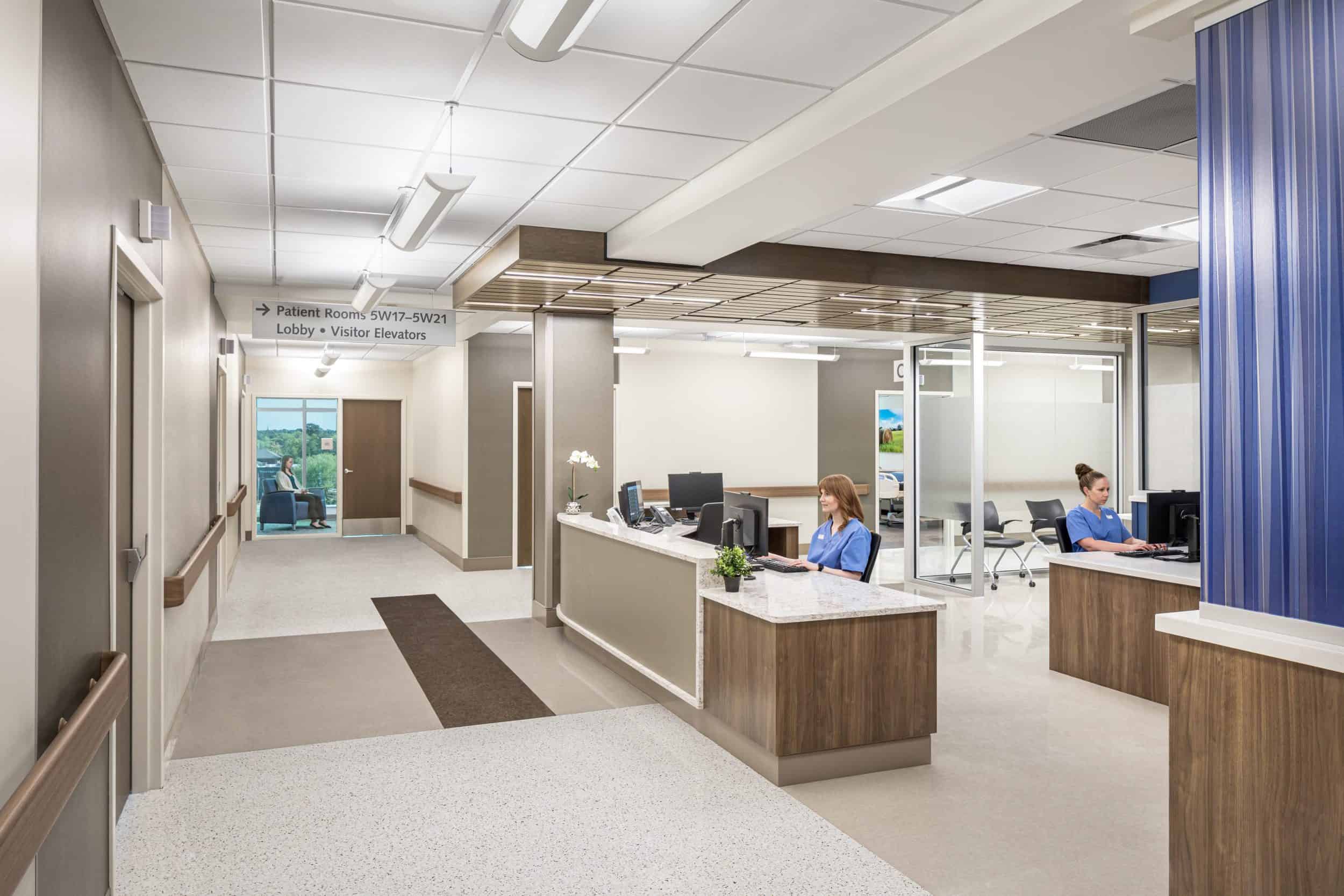 The image shows a nurses' station or reception area in the WakeMed Cary Hospital. There's a main reception desk with a wooden base and light-colored countertop where a staff member in blue scrubs is working at a computer. To the right, another staff member in blue is at a separate workstation. Above the hallway, there's a directional sign that reads 