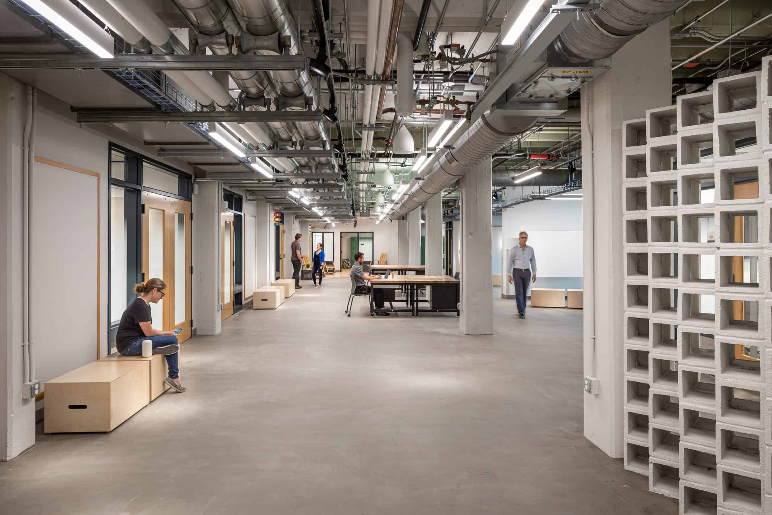 This image shows an industrial-style office hallway or collaborative workspace with exposed ceiling infrastructure. The space features a polished concrete floor and white walls with various doors and openings along the sides. Several people are visible - one person sitting on a wooden storage bench on the left, another at a desk or table in the middle, and a man in a light shirt walking down the corridor. The ceiling reveals exposed ductwork, pipes, and lighting fixtures, creating an urban loft aesthetic. On the right side, there's a decorative concrete block wall with a grid pattern allowing light to filter through. The minimalist design combines functional elements with modern workspace concepts, suggesting this may be part of a converted industrial building now serving as a creative or tech office environment.