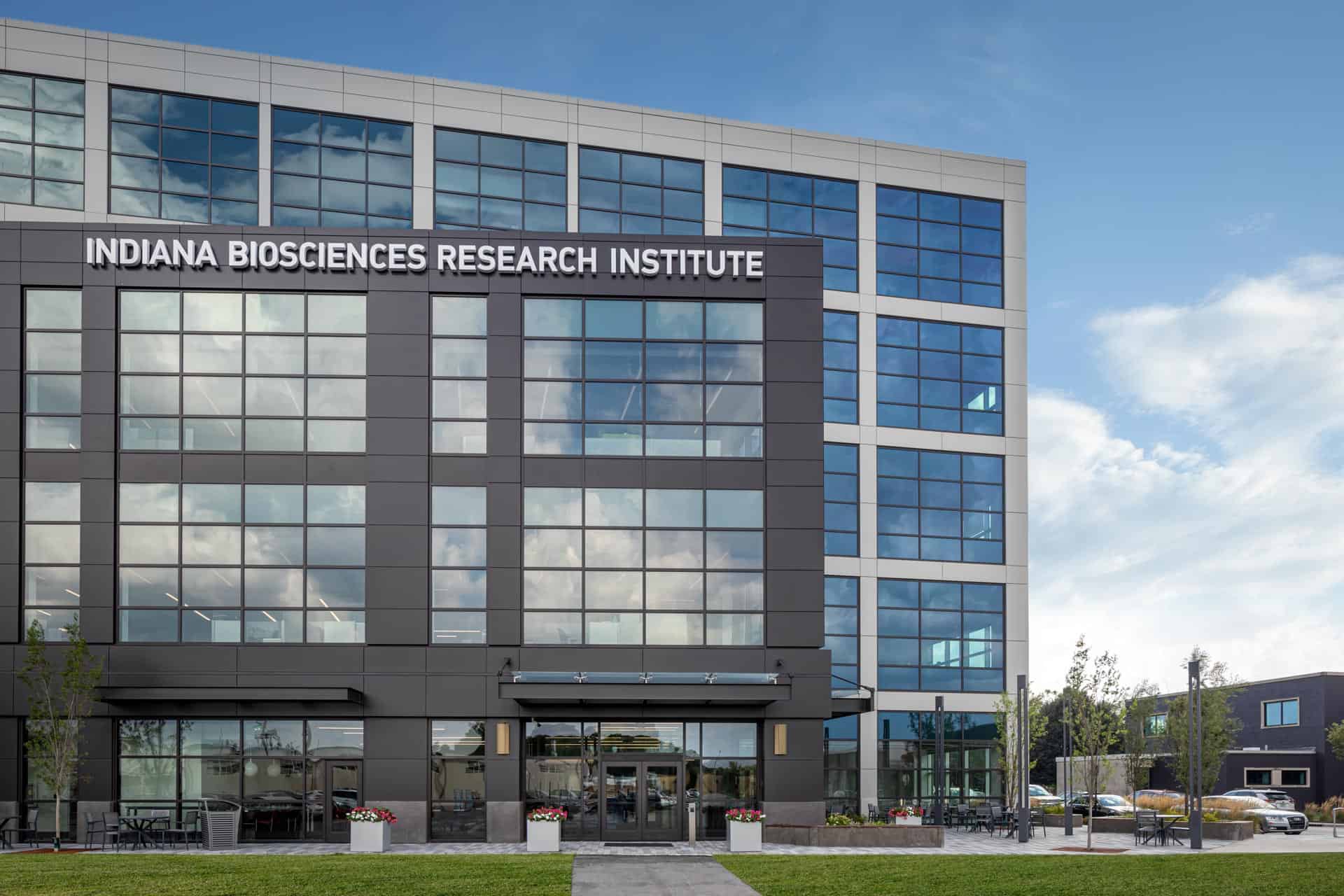 The image shows the exterior of the Indiana Biosciences Research Institute building. It's a modern, multi-story structure with a predominantly glass facade that reflects the sky and clouds. The building features a contrast of dark gray and light gray/white cladding materials, with large windows arranged in a grid pattern across the facade. At the top of the main entrance area, there's prominent signage reading