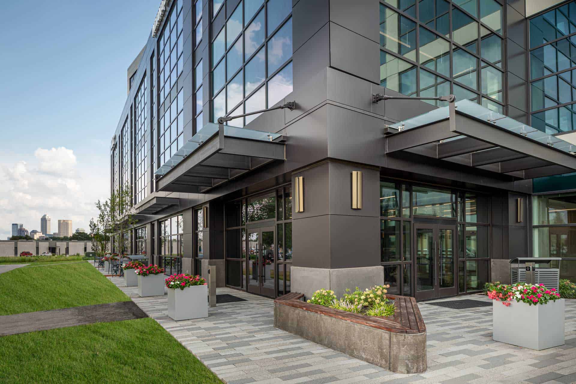 The image shows a close up of the exterior of the Indiana Biosciences Research Institute building. It's a modern, multi-story structure made up of mostly uniform windows. The main entrance has glass doors with a small canopy overhead and appears to have some planters with flowers flanking the entrance.