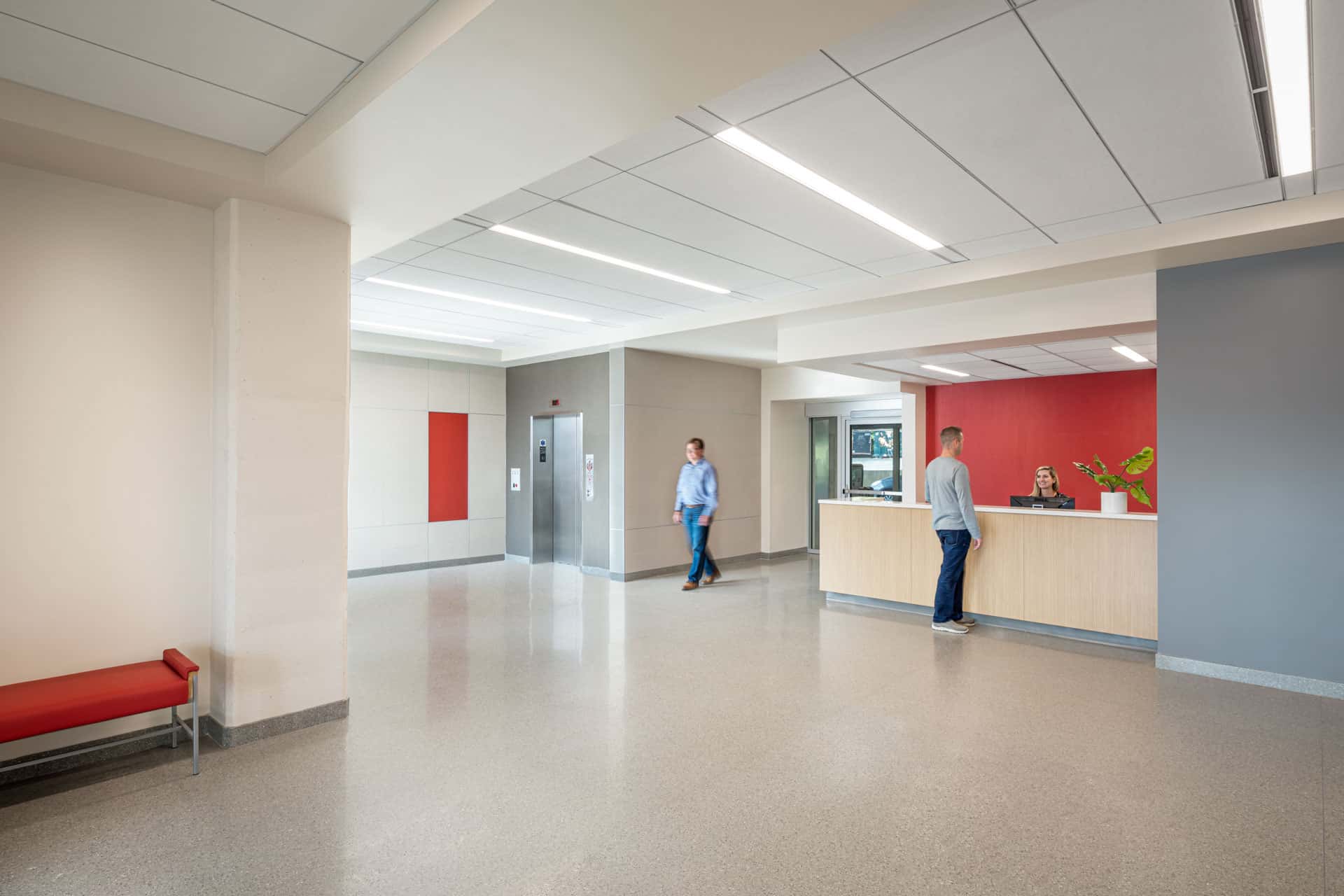 This image shows a modern reception area for the Luddy Center for Artificial Intelligence. The light oak reception desk stands out amongst the rest of space because behind it is a bright red wall while the rest of the walls are neutral-colored and minimalistic. There is a single elevator to the left of the reception desk. A woman is seated behind the reception desk and talking to a man, likely checking him into the building or providing directions. There is a modern red bench in the lobby, complementing the red accent wall. The space has contemporary design elements including recessed linear lighting, a drop ceiling, polished floors, and no decorations aside from one plant on the reception desk.