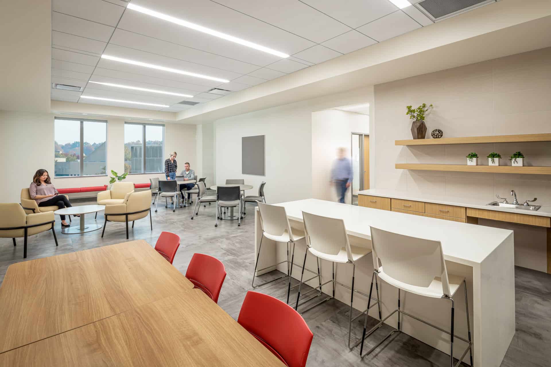 This image shows a modern collaboration space or break room inside the Luddy Center for Artificial Intelligence. The room features a small kitchenette on the right side with light oak cabinets, white countertops, and an island bar with white chairs. At the very front of the image is a table made of the same light oak with red chairs, possibly an area for lunch or open meetings. In the background are more configurations of beige accent chairs and grey tables with matching grey chairs. The space has contemporary design elements including recessed linear lighting, polished concrete floors, and clean neutral-colored walls.
