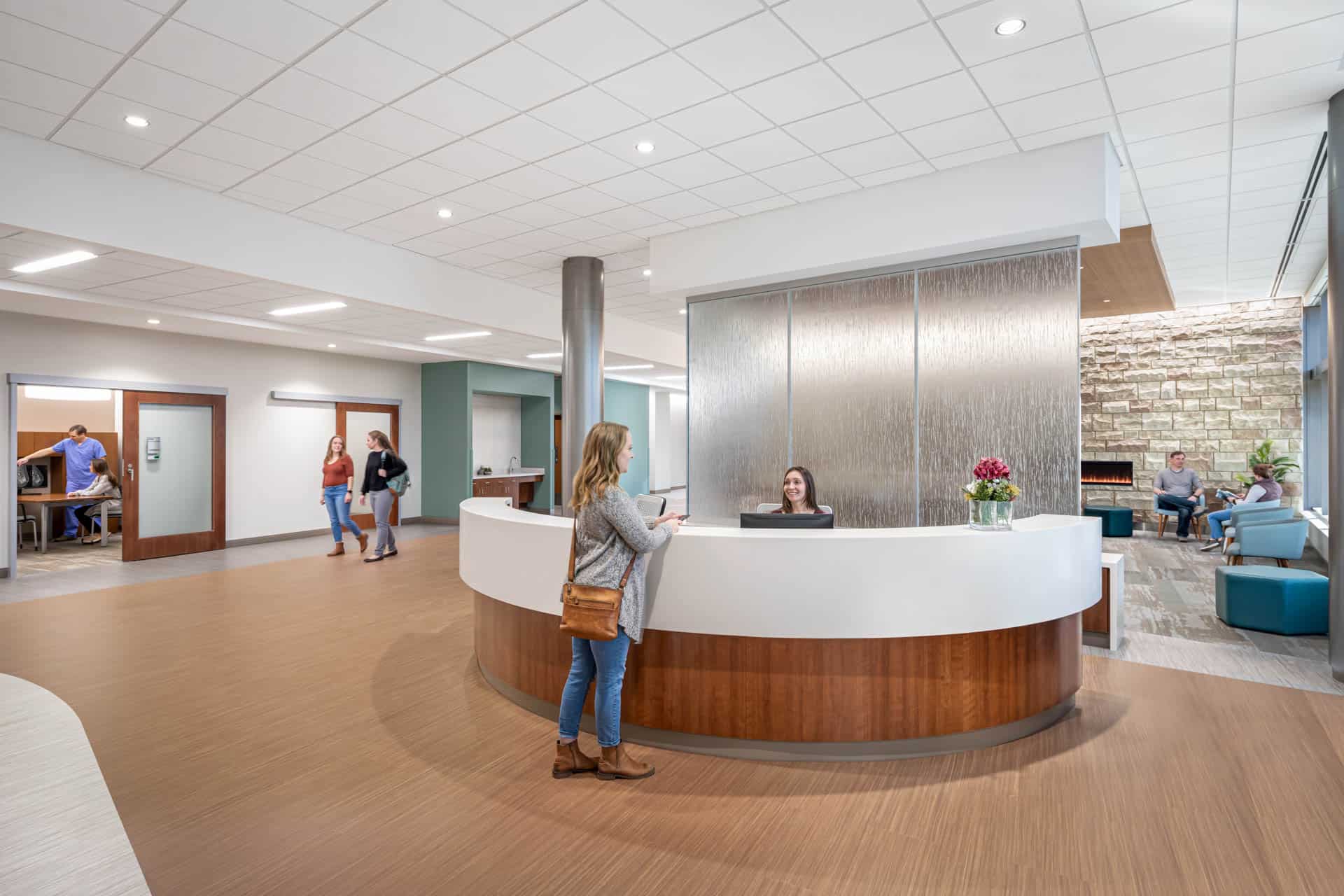 Modern healthcare reception area with a curved white and wood-toned welcome desk. A woman in a grey top and jeans stands near the desk, with a receptionist seated behind. Behind the desk is a textured glass pall separating the reception space from the waiting room while maintaining an open feel. The waiting space behind the desk features a stone accent wall with colorful abstract artwork and a linear fireplace. The room has a bright, open design with recessed lighting and a clean, welcoming aesthetic. This image shows BSA's expertise in designing welcoming spaces that enhance the patient experience.