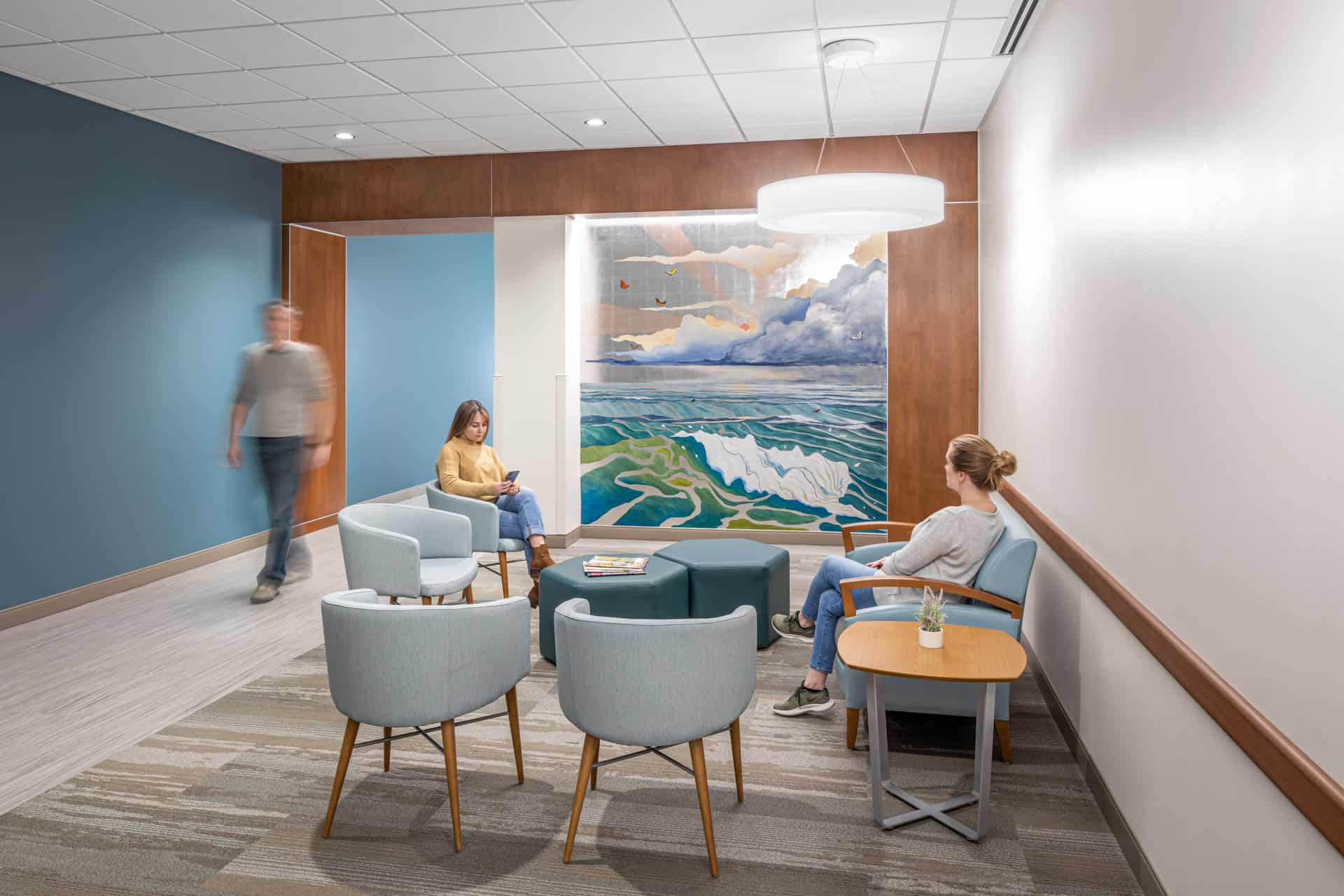 A modern enclosed interior patient waiting room of the UNC Rex Cancer Center centered around an abstract mural of an ocean. The space is furnished with light blue and green seating including chairs and ottomans. Two people are seated in the lounge, one scrolling on her phone.
