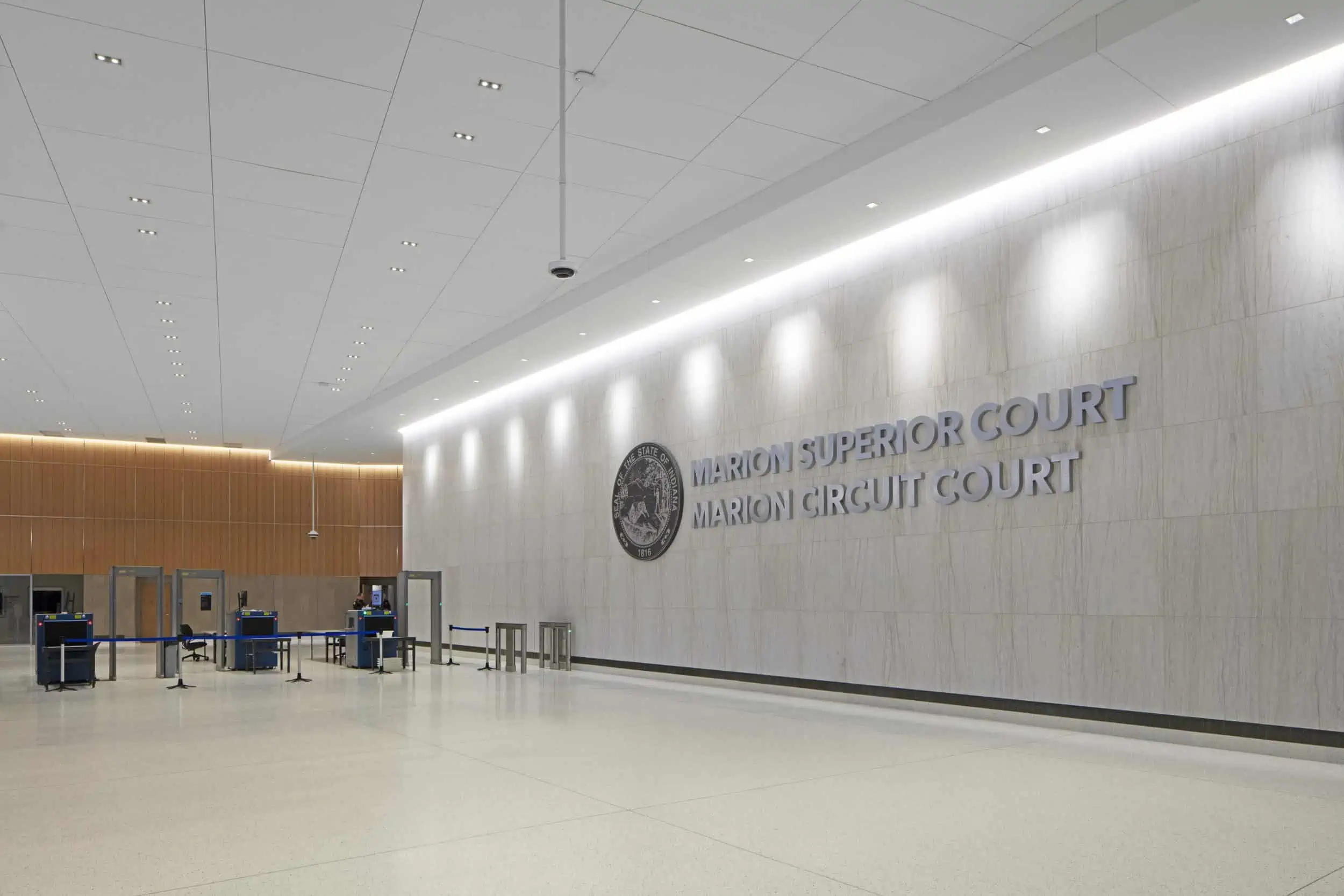 The image shows the interior lobby of the Marion Superior Court and Marion Circuit Court. The space features a minimalist, modern design with a large white marble or stone wall that prominently displays the court names in silver metallic lettering alongside what appears to be an official seal or emblem. The ceiling is white with recessed lighting, and there's elegant ambient lighting along the wall. To the left side of the image, there's a security checkpoint area with metal detectors and screening stations, typical of courthouse entry procedures. The floor is polished light-colored stone or tile, and the overall aesthetic is clean, spacious, and formal, befitting a judicial building. The design combines functionality with a sense of dignity appropriate for a courthouse.