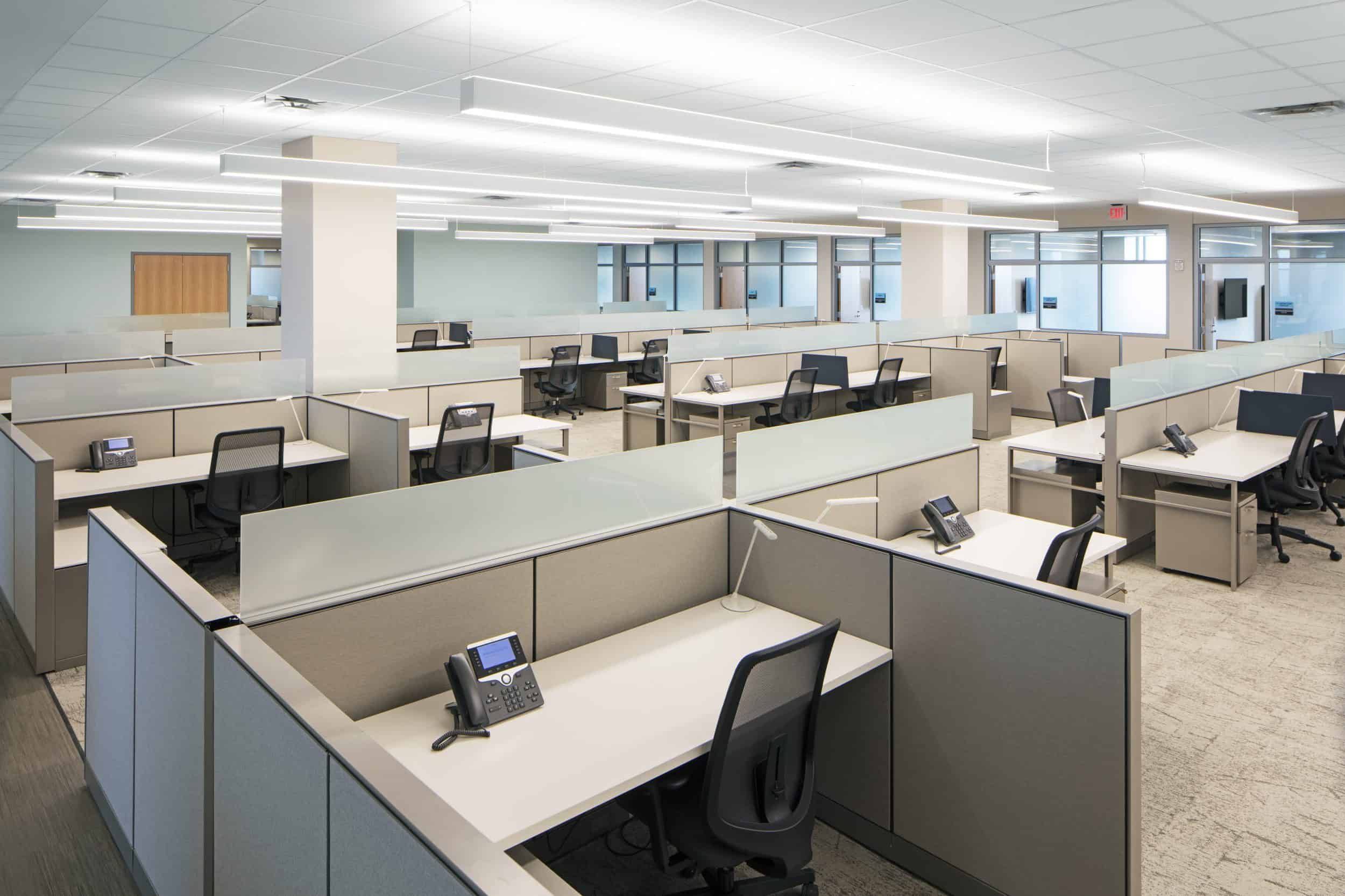 The image shows a modern office space with a standard cubicle layout. The open-plan area features numerous workstations arranged in a grid pattern, each separated by light-colored partition walls of medium height. Each cubicle is equipped with a desk, black office chair, and desk phone. The office has a clean, minimalist design with a neutral color palette predominantly featuring beige, white, and gray tones. The space appears to be well-lit with long fluorescent ceiling lights and includes some natural light from windows visible along the back wall. Support columns are integrated throughout the space, and the ceiling has a standard drop ceiling design with white panels. The floor appears to be commercial carpet in a light neutral tone. This appears to be a typical corporate or government administrative office environment designed for clerical or administrative staff, with workstations that provide some privacy while maintaining an open concept.