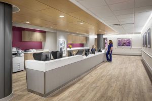 This image shows a modern nurses' station at Riley Hospital for Children at IU Health Maternity Tower featuring a curved design with a distinctive wood-toned ceiling element that follows the station's contours. The workspace includes a long white countertop with computer workstations where staff members are seated, complemented by light wood cabinetry and burgundy accent walls as seen in patient rooms. Two healthcare workers in blue scrubs are visible - one seated at the workstation and another standing nearby. The space features contemporary finishes including wood-look flooring, and the corridor walls display framed artwork including images of purple cherry blossoms, creating a welcoming and professional healthcare environment.