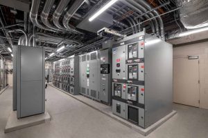 This image shows the electrical switchgear room at Riley Hospital for Children at IU Health Maternity Tower, featuring a row of large gray electrical distribution panels and control equipment. The room contains multiple electrical switchgear cabinets with various meters, switches, and control interfaces that manage the hospital's power distribution systems. An extensive network of conduits, pipes, and ductwork runs along the exposed ceiling, typical of a hospital's critical infrastructure areas. The space has concrete flooring with proper electrical grounding and safety measures, and includes emergency lighting systems. This electrical room is essential for providing reliable power distribution throughout the hospital facility, ensuring continuous operation of medical equipment and building systems vital to patient care.